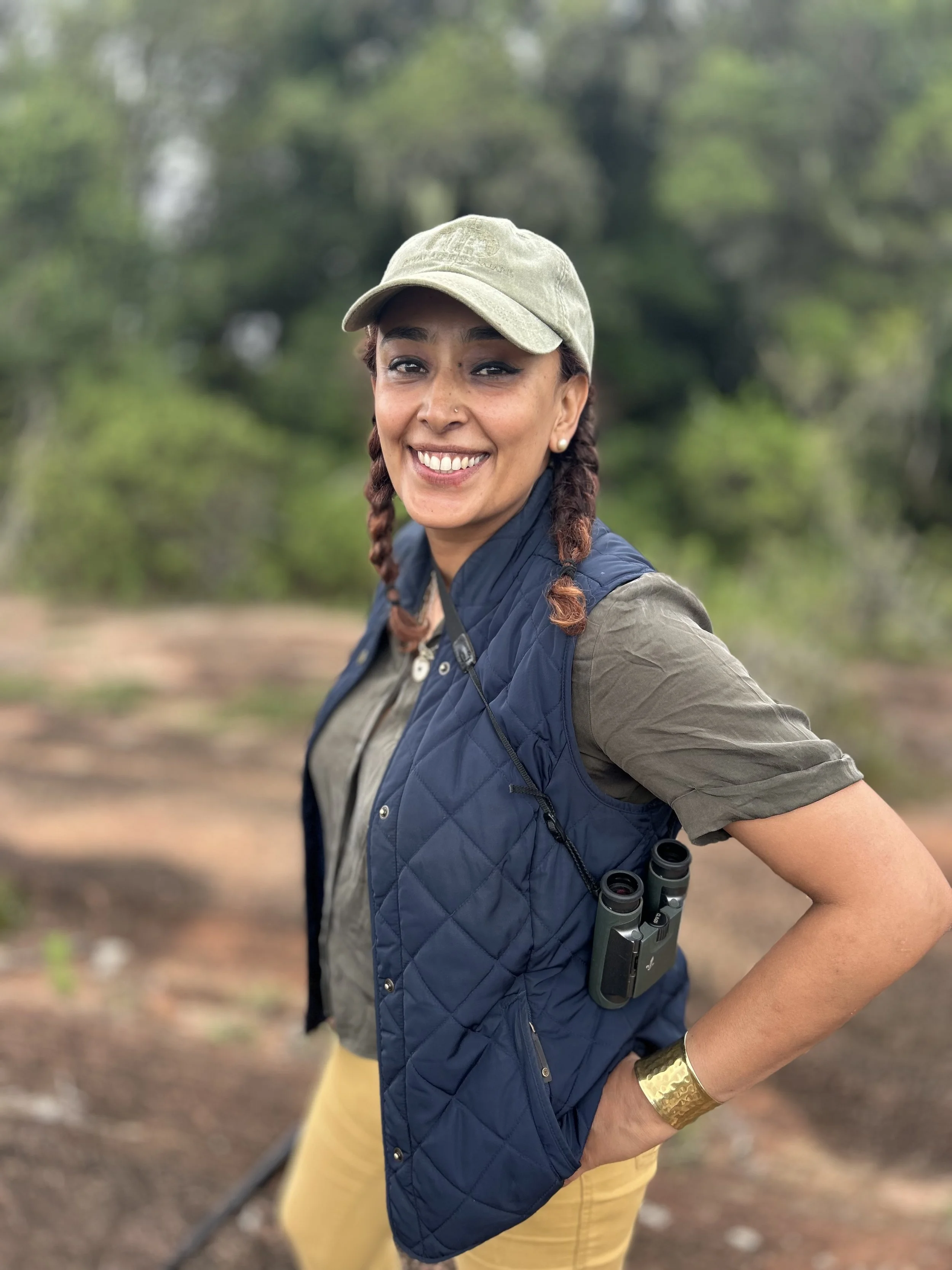 Smiling woman outdoors wearing a beige baseball cap, navy quilted vest, and yellow pants, with binoculars hanging from her vest, in a green, wooded area.