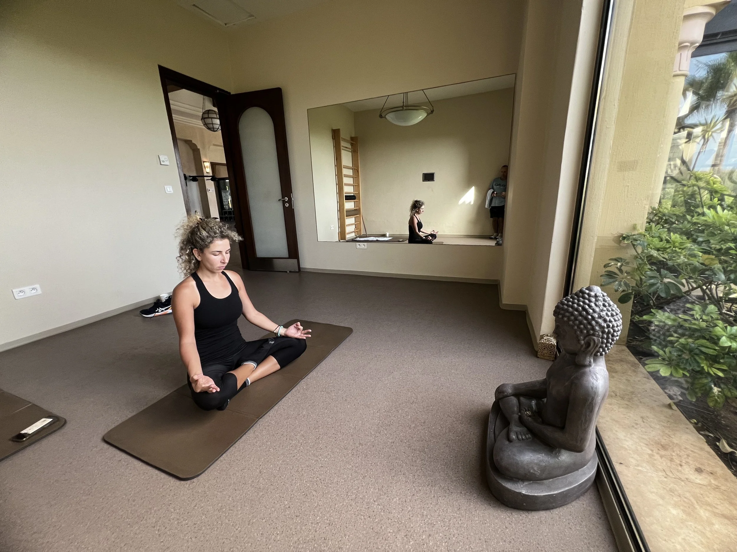 Three women practicing yoga indoors, one sitting in a meditative pose on a yoga mat, with large windows showing greenery outside.