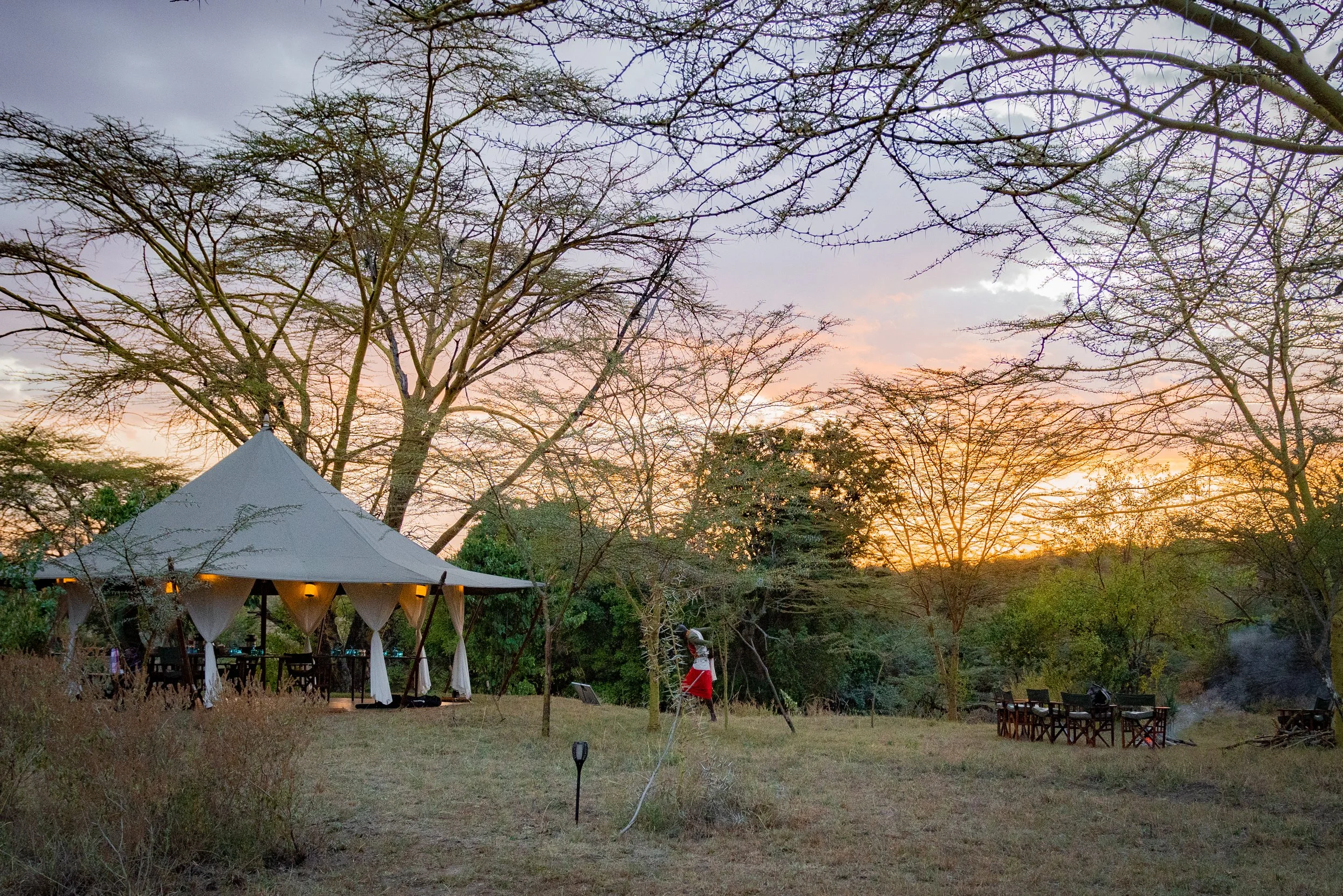An outdoor scene at sunset with a large tree and a gazebo with a pointed roof. There are string lights inside the gazebo, and a person dressed in red and another person are visible nearby. There are additional chairs and tables under a tree to the ri