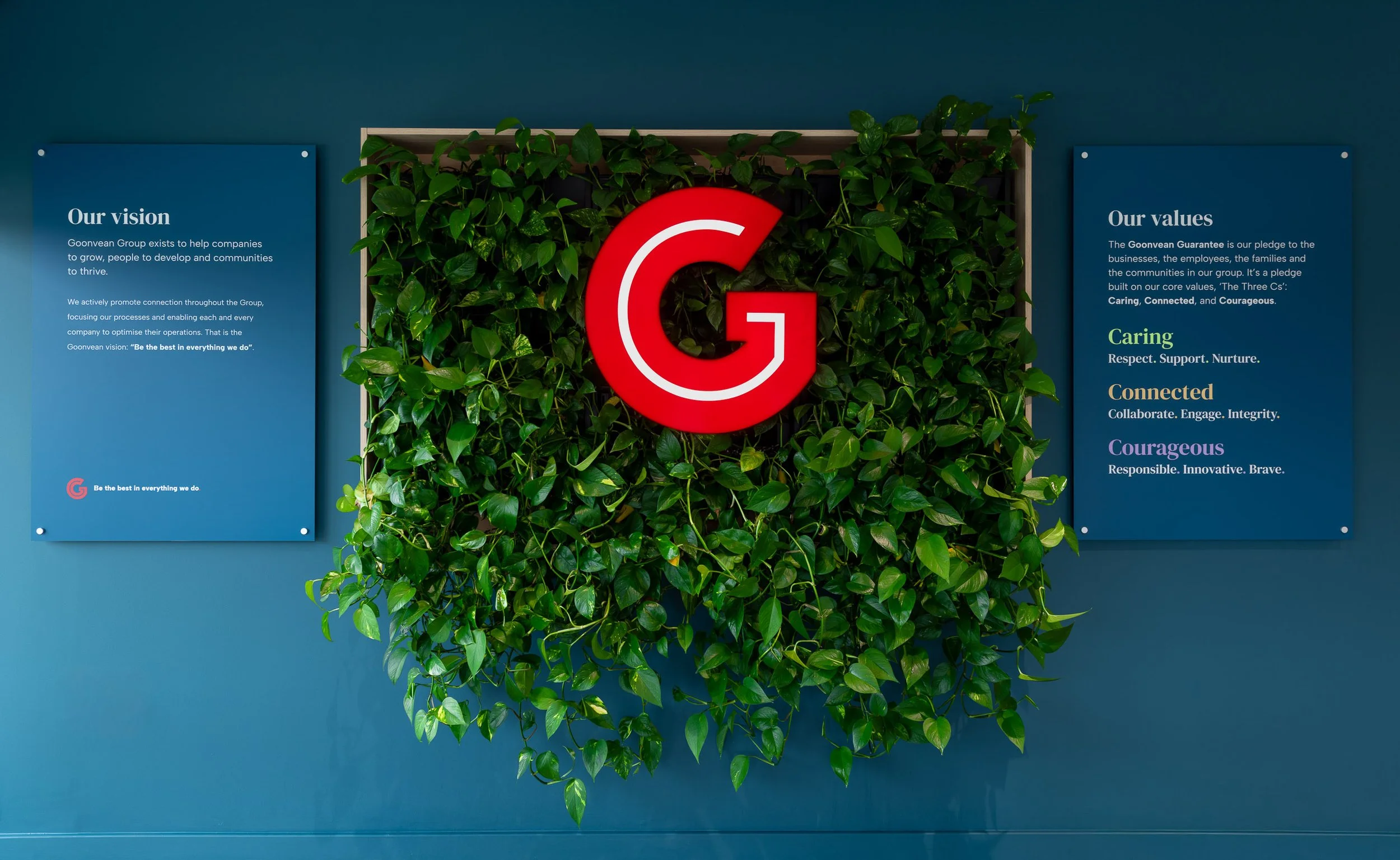 A wall with green leafy plants and three blue informational panels. The center panel has a large red G logo. The left panel discusses the company's vision, and the right panel lists core values including Caring, Connected, and Courageous.