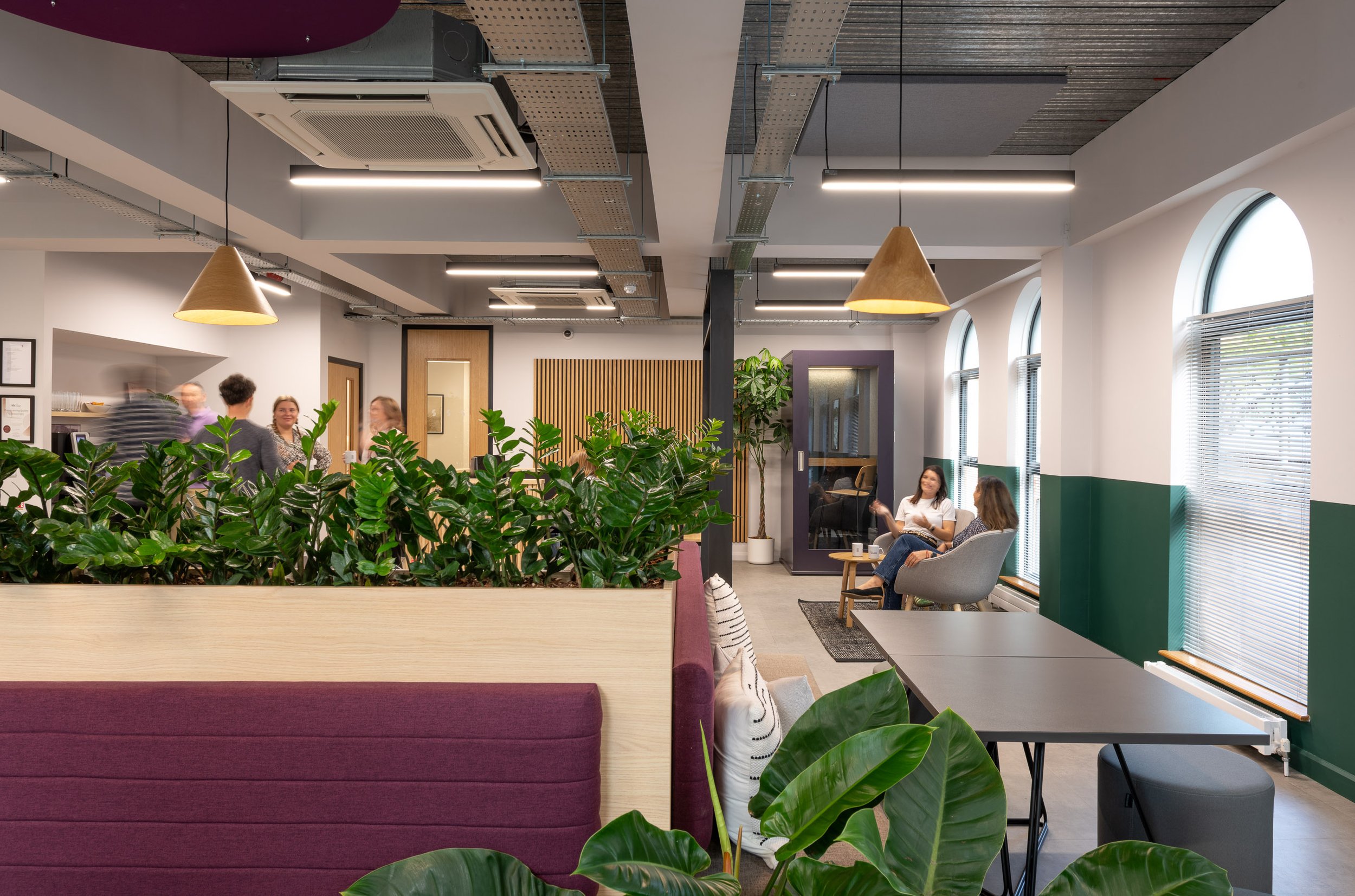 Interior of a modern office lounge area with large windows, green plants, people sitting and socializing, wooden furniture, pendant lighting, and an air conditioning unit on the ceiling.