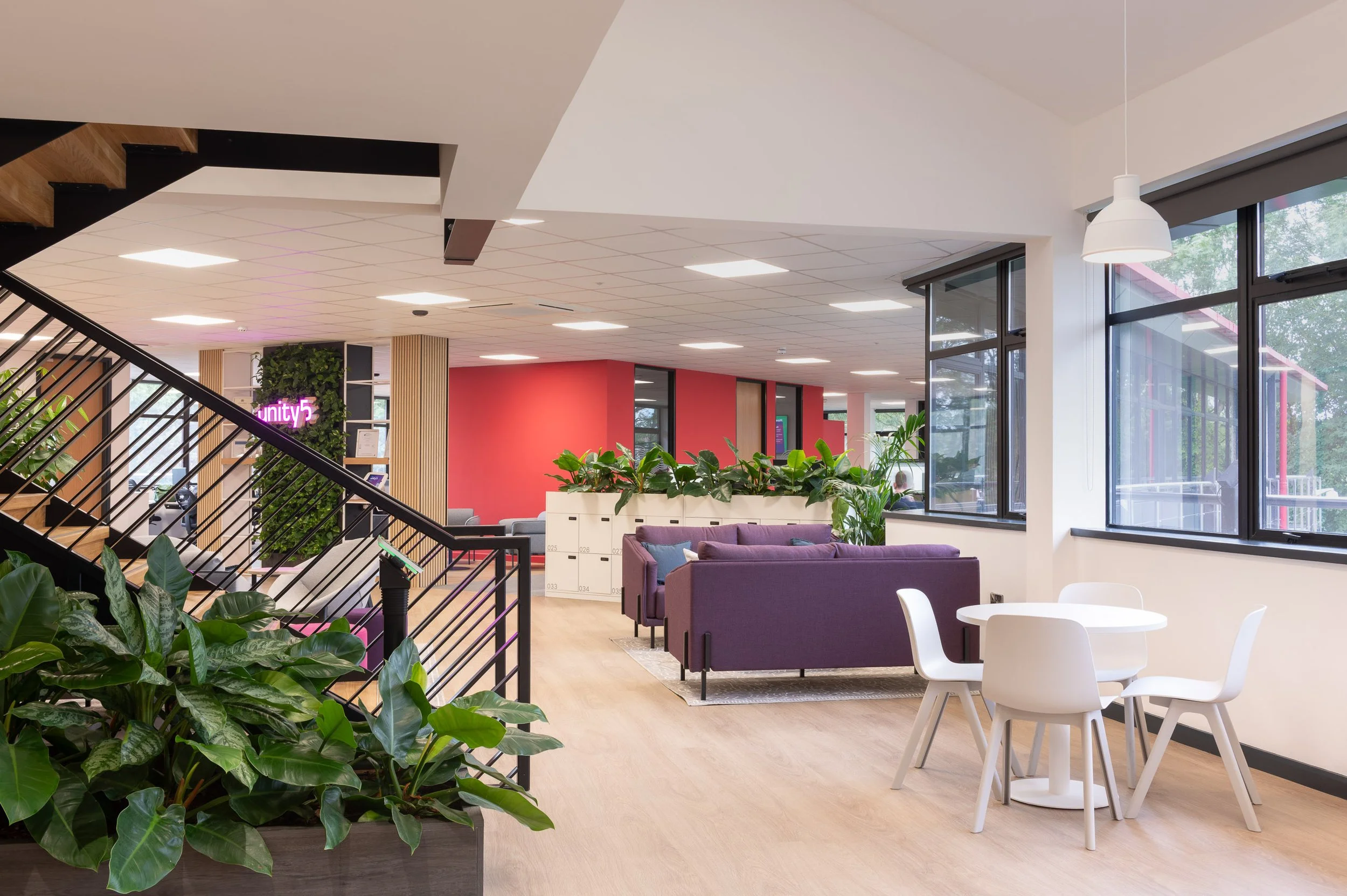 Interior view of a modern office or coworking space with seating area, plants, large windows, and a staircase with black railings.