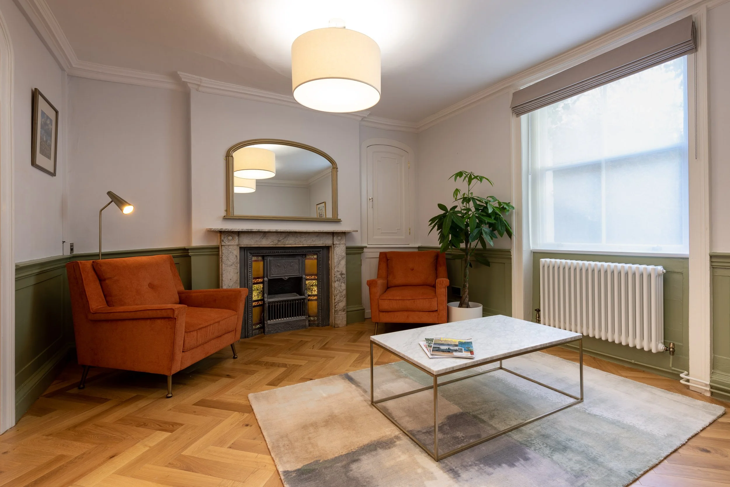 Living room with orange armchairs, a marble fireplace with a gold-framed mirror, a potted plant, a coffee table with magazines, large window, ceiling light, wooden flooring, and wall art.