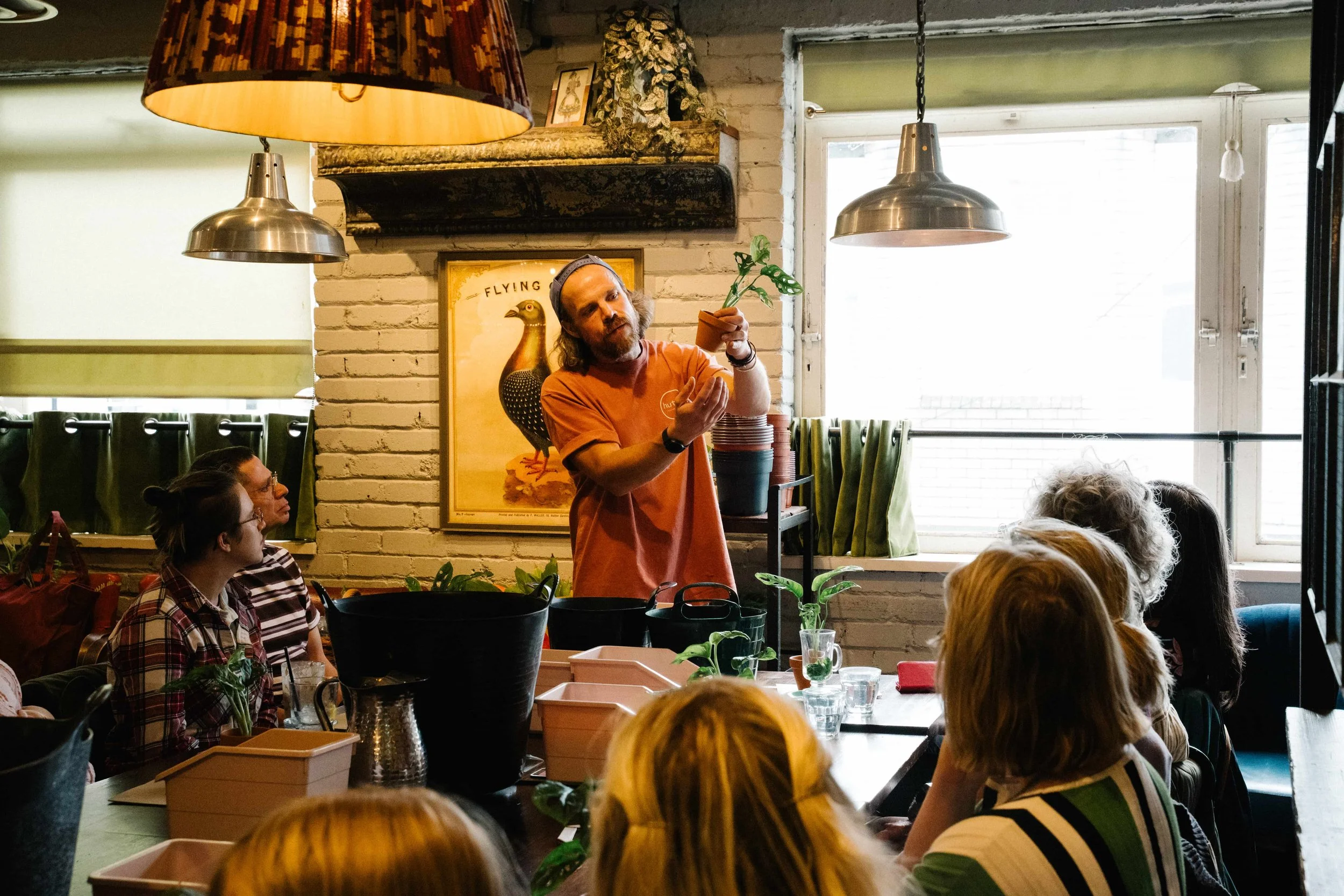 Man with long hair and beard, wearing an orange T-shirt, stands at a table holding a small terracotta pot with green plants inside, engaging with seated people in a cozy, well-lit room with large windows, plants, and vintage decor.
