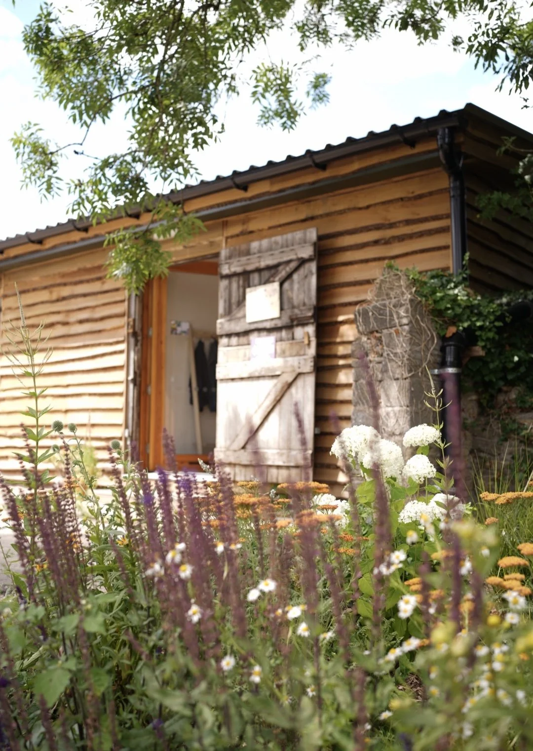 A rustic wooden shed with a slightly open sliding door, surrounded by blooming wildflowers and garden plants, with a tree overhead and a stone sculpture to the right.