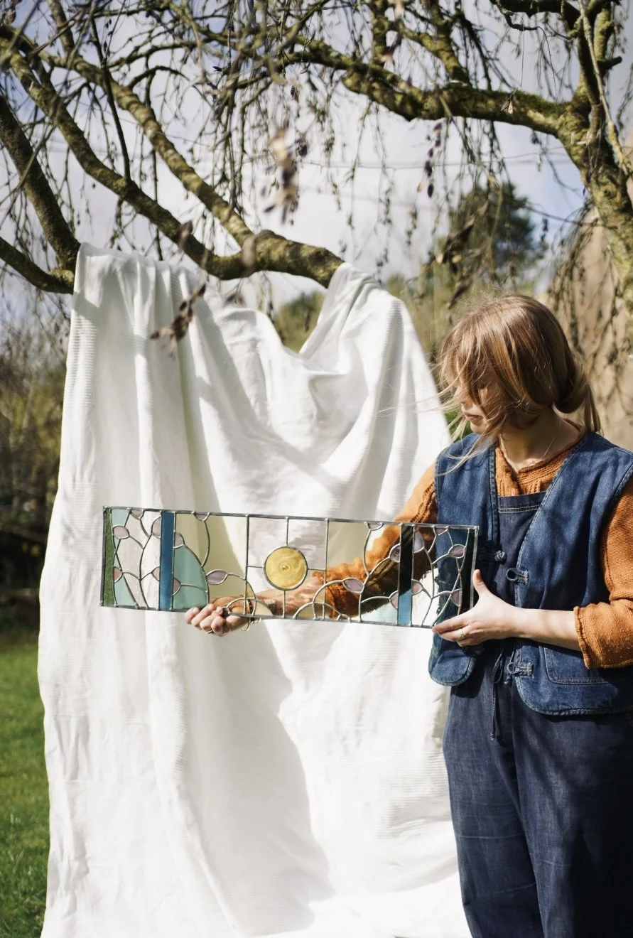 A woman with shoulder-length brown hair, wearing a denim vest over a rust-colored sweater, holding a stained glass panel outdoors near a white sheet hanging from tree branches.