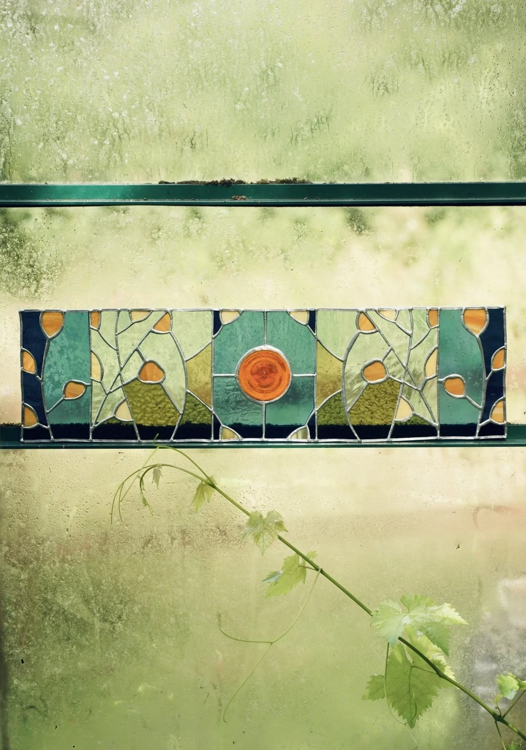 Close-up of a decorative stained glass panel with abstract leaf and vine patterns and a central orange circle, set against a foggy, greenish background with a plant vine in the foreground.