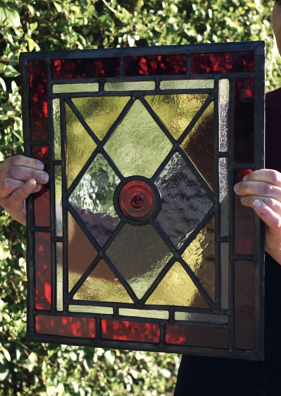 Person holding a stained glass window panel outdoors with green foliage in the background.