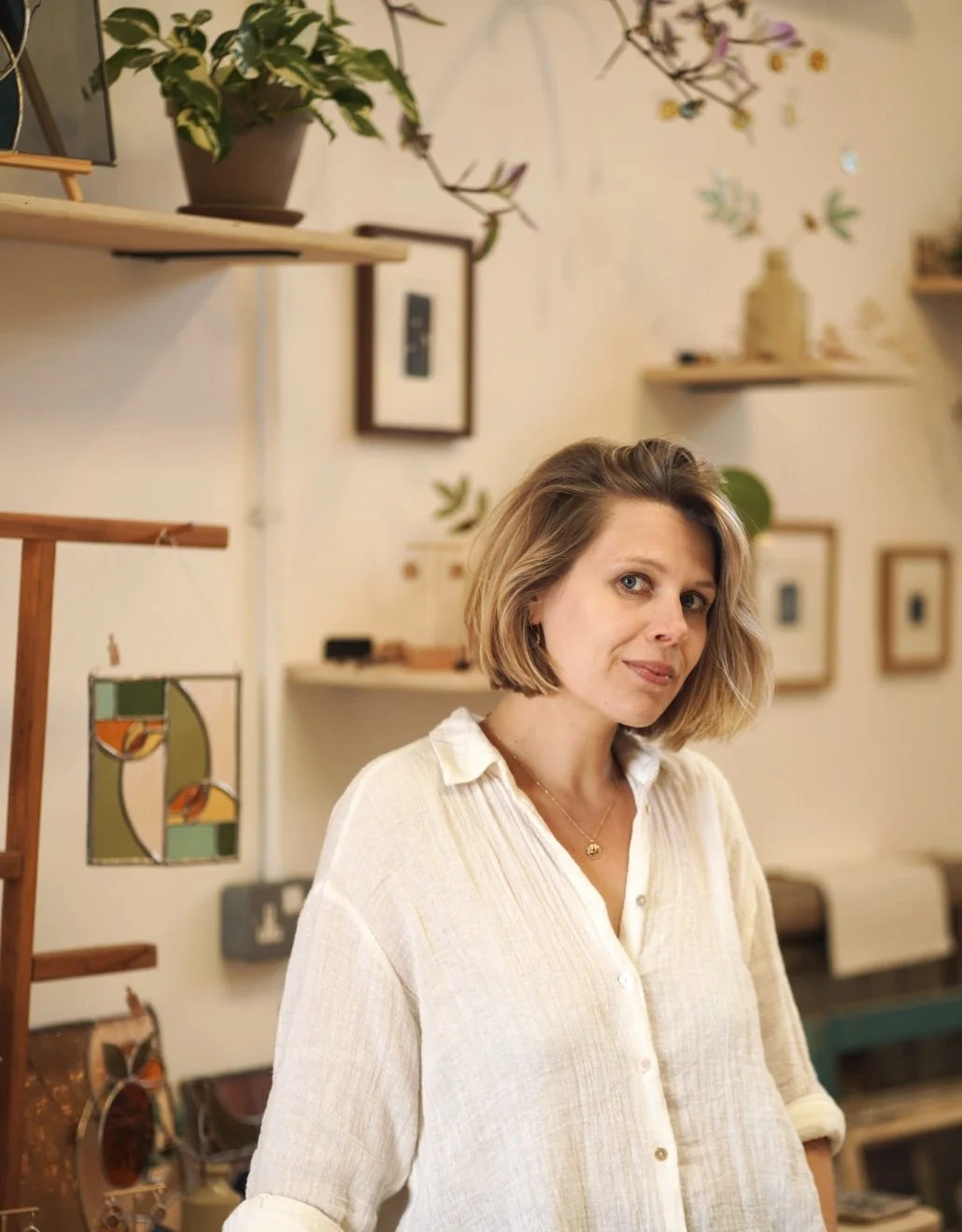 A woman with short blonde hair wearing a white shirt stands in an art gallery or studio with framed artwork and decorative items on shelves and walls behind her.