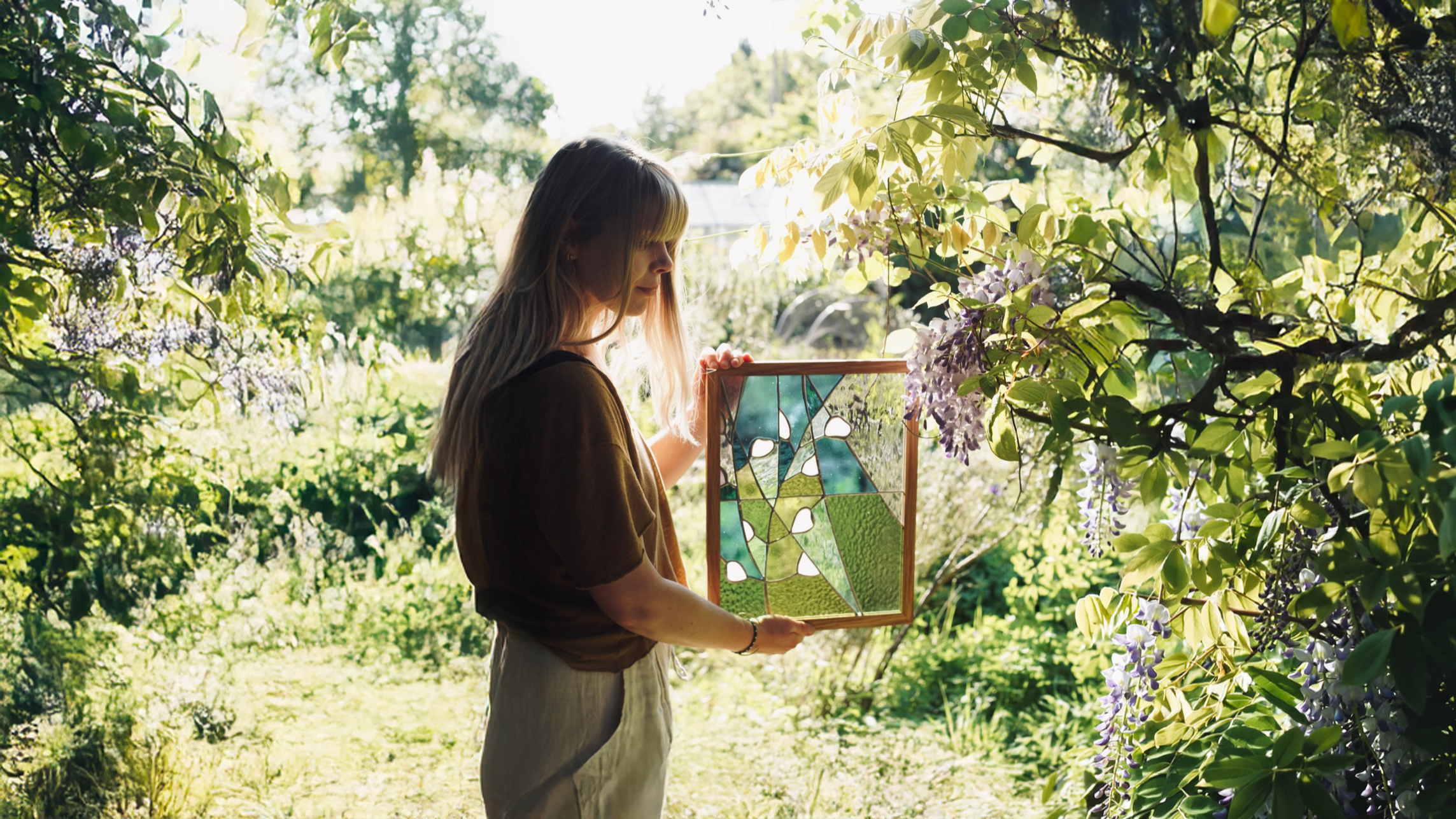 A woman with long hair holding a stained glass window while standing among lush green trees and flowering plants in bright sunlight.