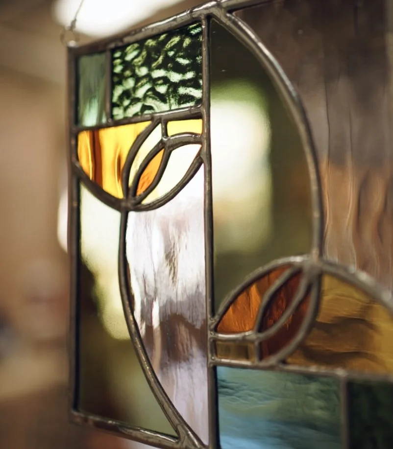 Close-up of a stained glass window with abstract geometric and floral patterns in green, amber, clear, and blue glass.