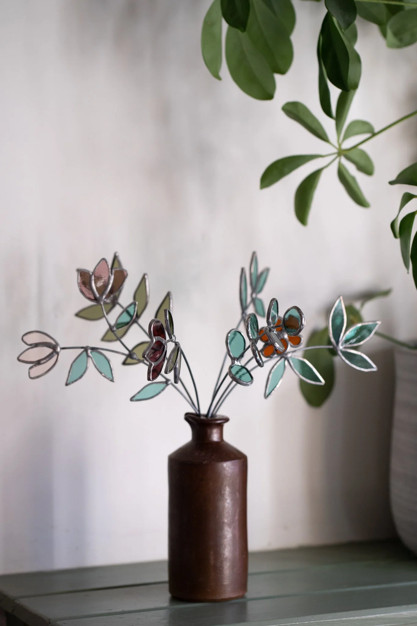 A brown ceramic vase holding an arrangement of decorative metal leaves with colored enamel accents, placed on a wooden surface with a plant with green leaves in the background.