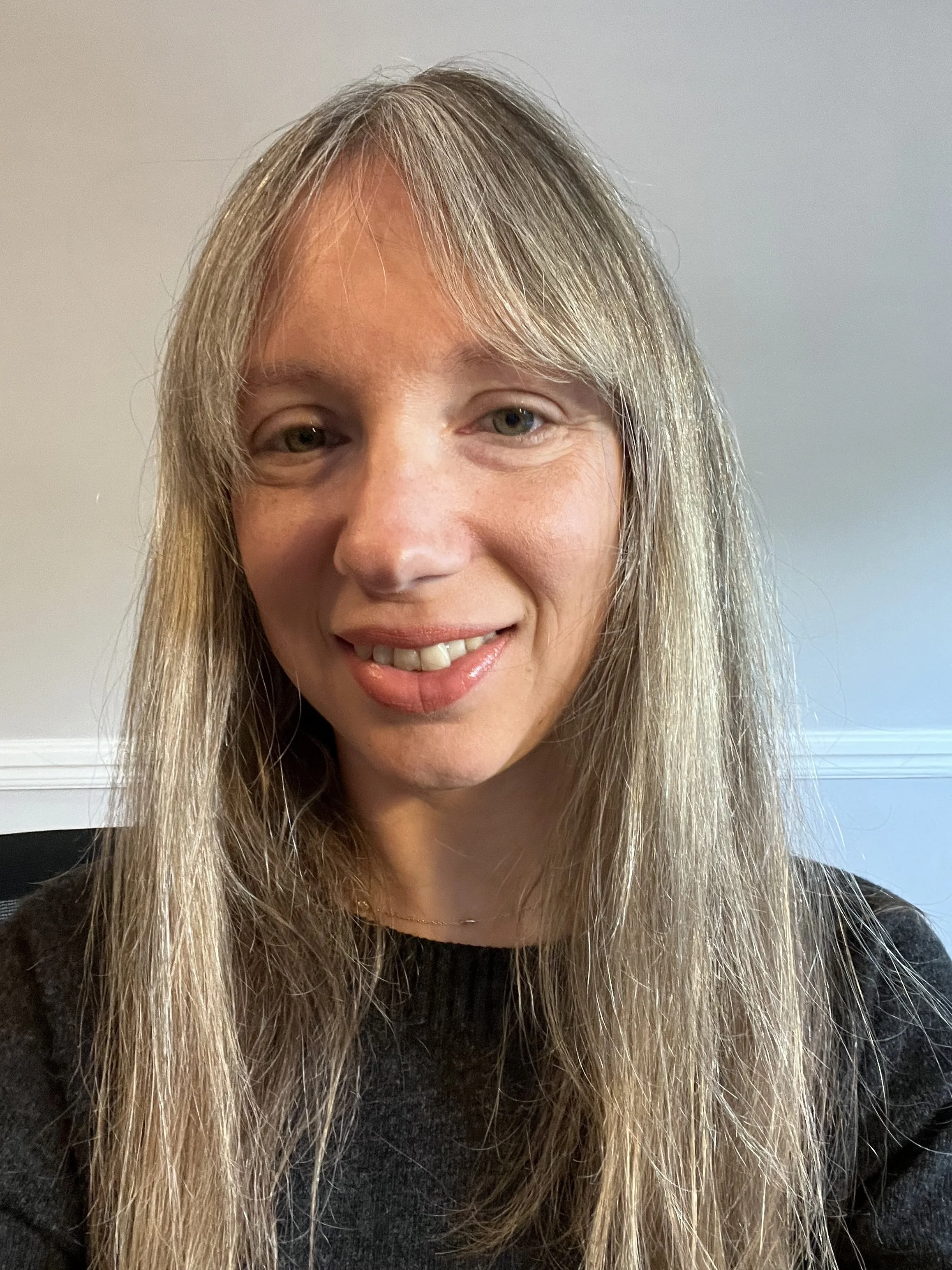 A woman with long blonde hair, smiling, wearing a black top, in front of a light-colored wall.