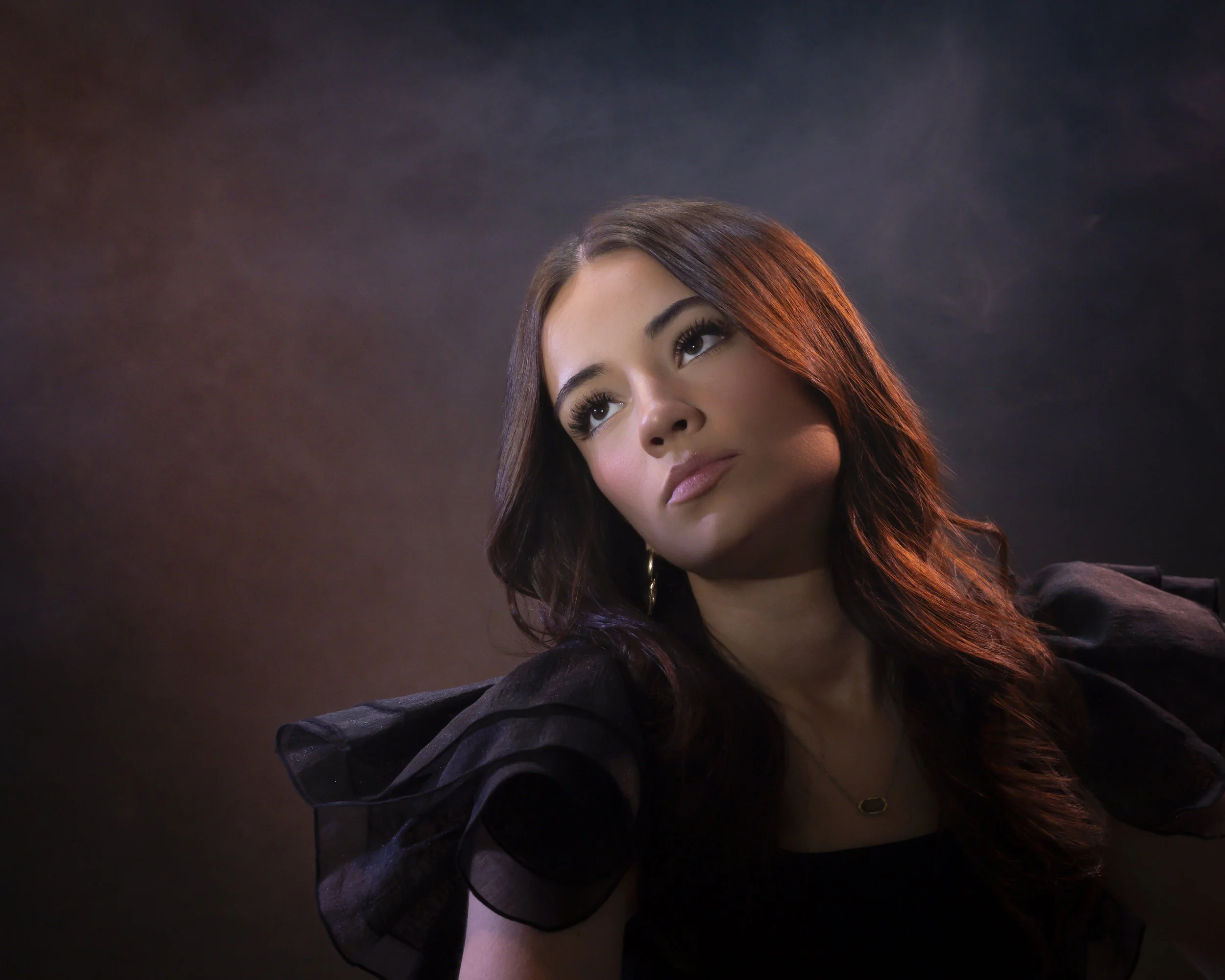 Teenage girl with long dark hair looking upward in a black top with puffed sleeves against a dark moody background