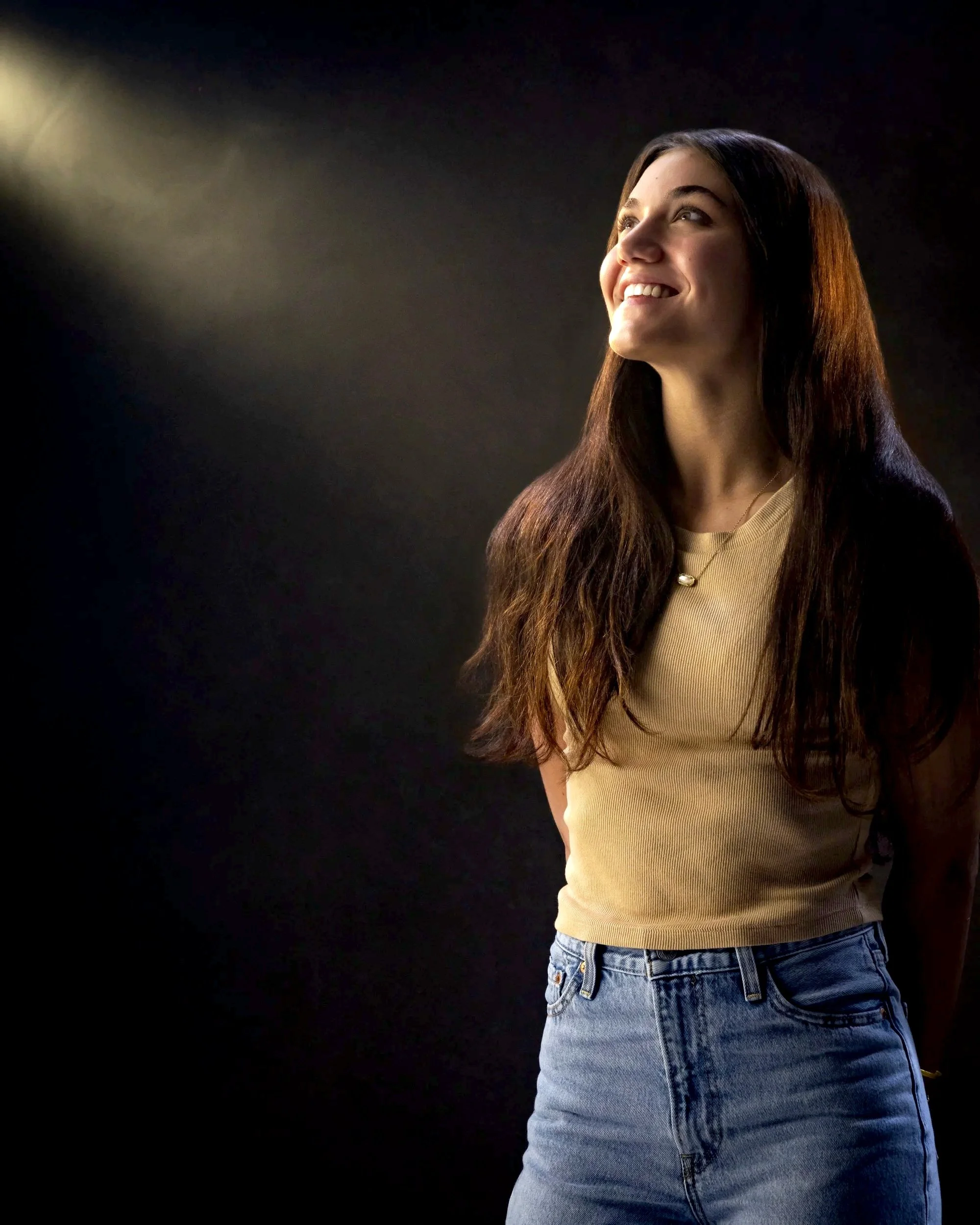 Smiling teenage girl in a beige top and jeans looking upward, lit by a beam of light from the left