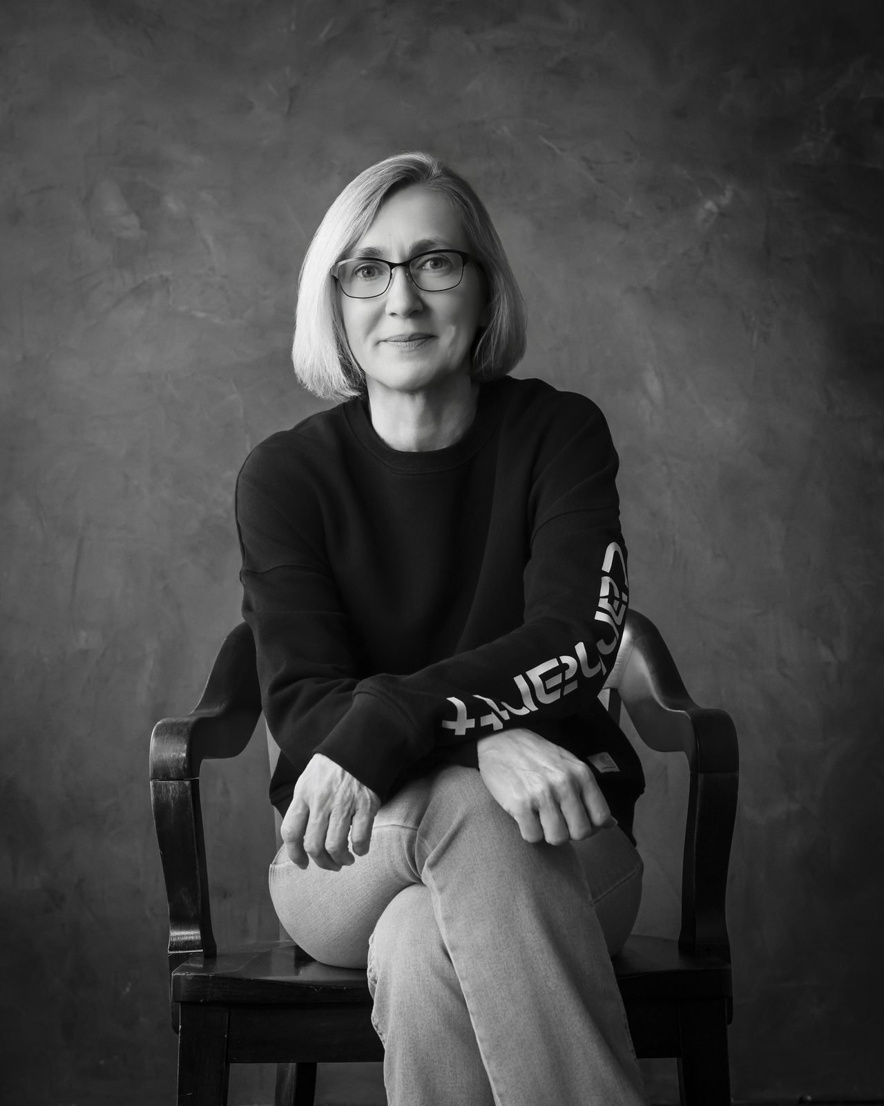 Black and white portrait of a woman with glasses seated on a wooden chair against a textured backdrop
