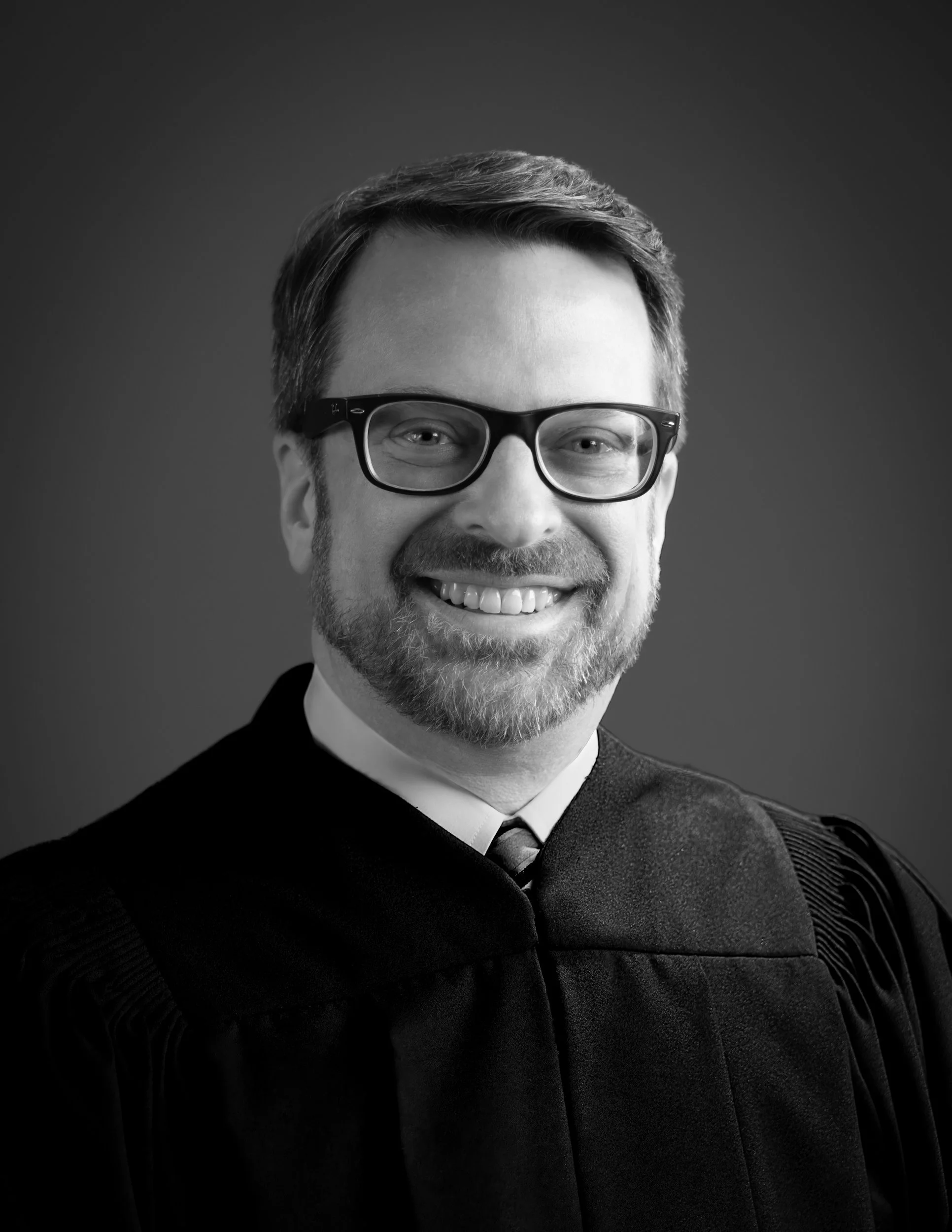 Professional black and white headshot of a smiling bearded man in glasses wearing a judge's robe