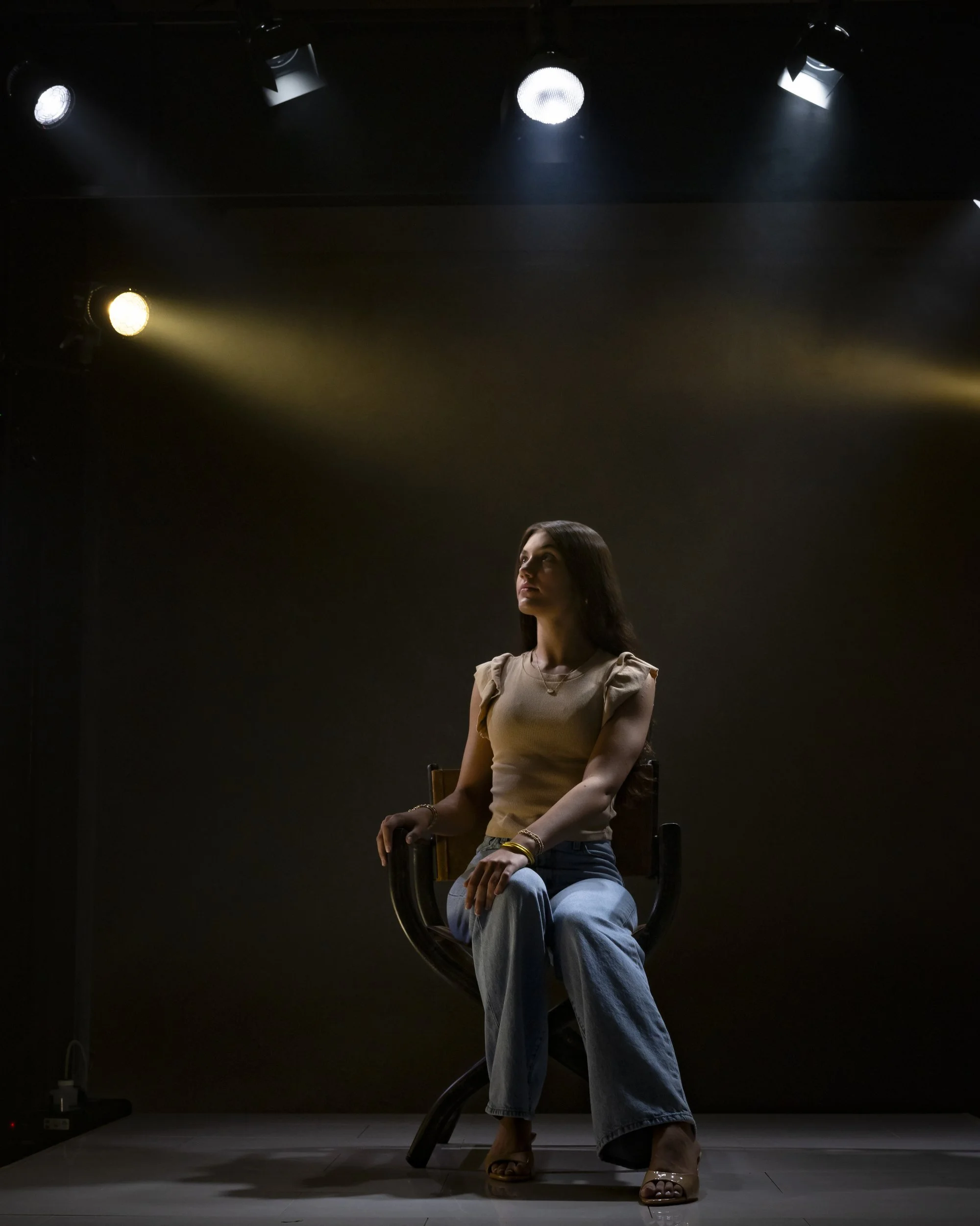 Teenage girl seated on a chair in a dimly lit studio with overhead spotlights, in a beige top and blue jeans