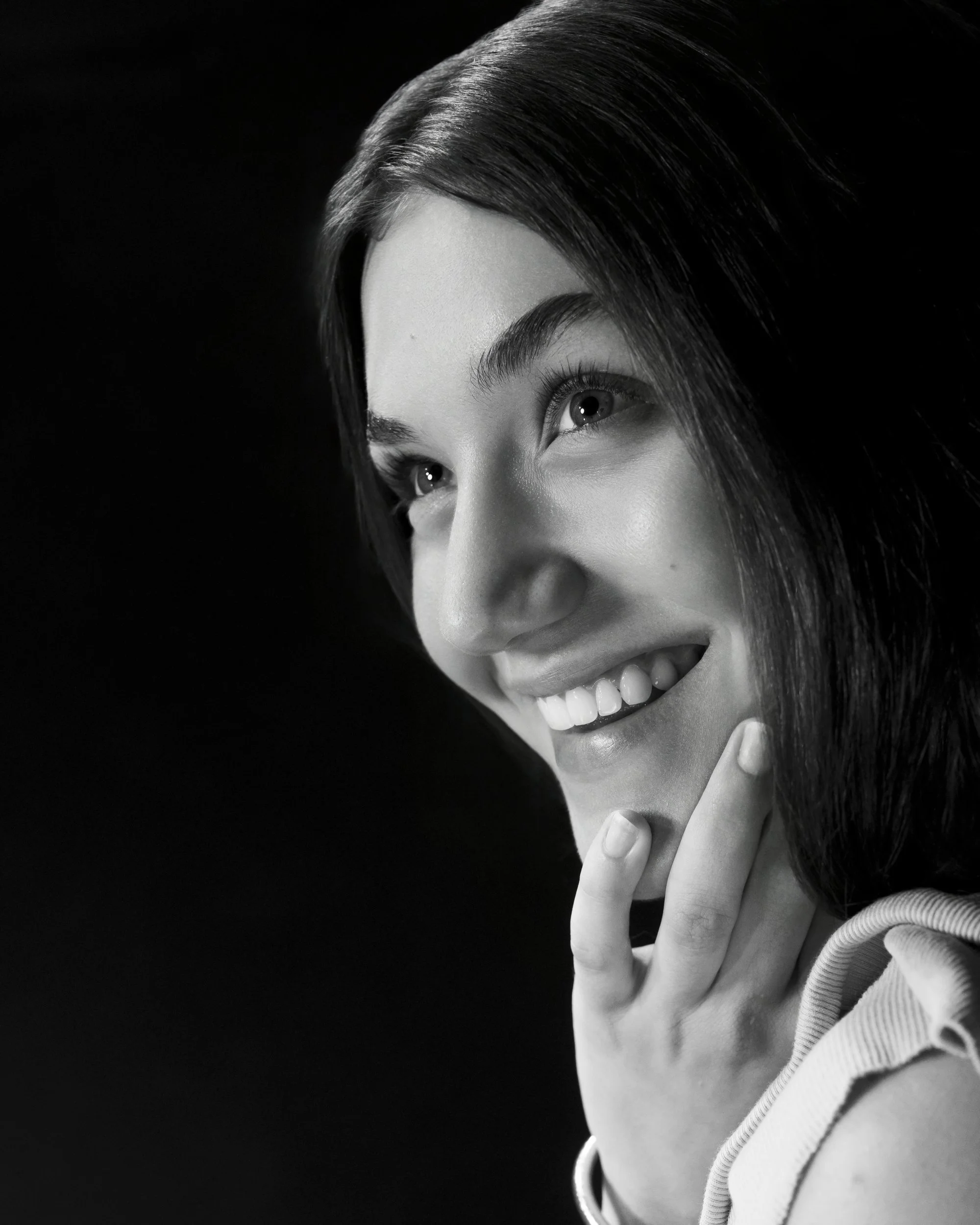 Close black and white portrait of a smiling teenage girl with her hand on her chin and bright eyes