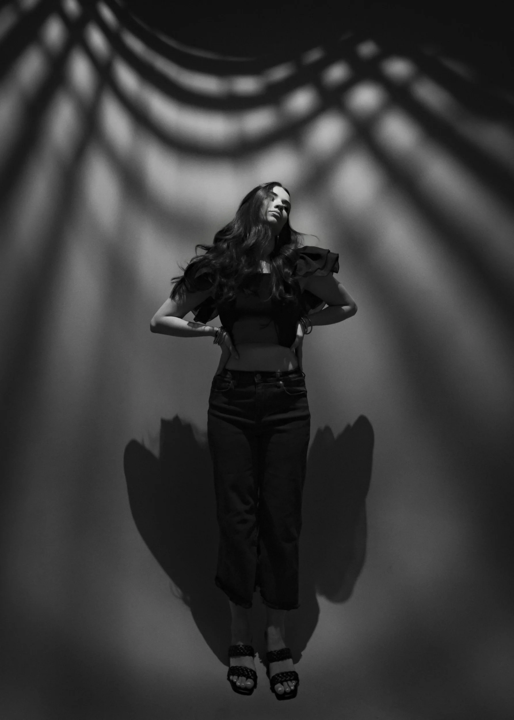 Black and white portrait of a teenage girl standing with her hands on her hips, patterned shadows on the wall behind her