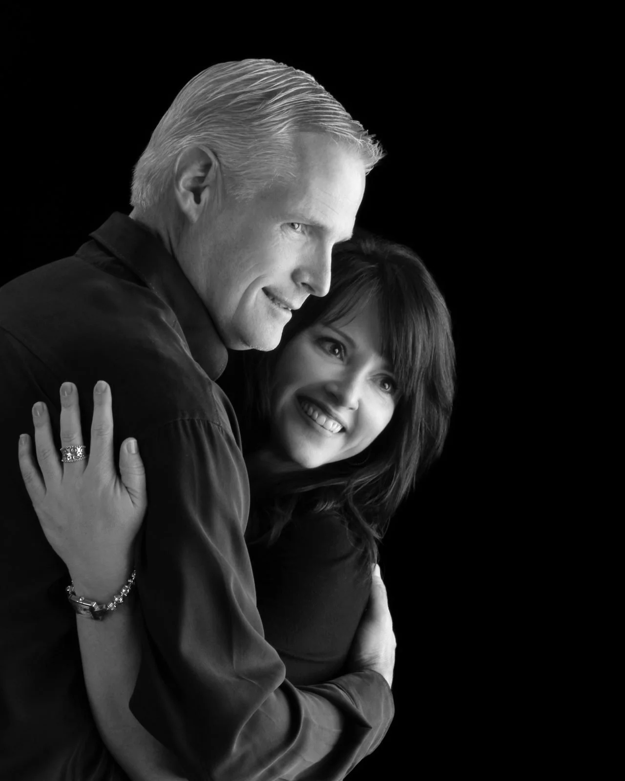 Black and white photo of two smiling people, a man with slicked-back gray hair and a woman with dark wavy hair, embracing each other against a black background.