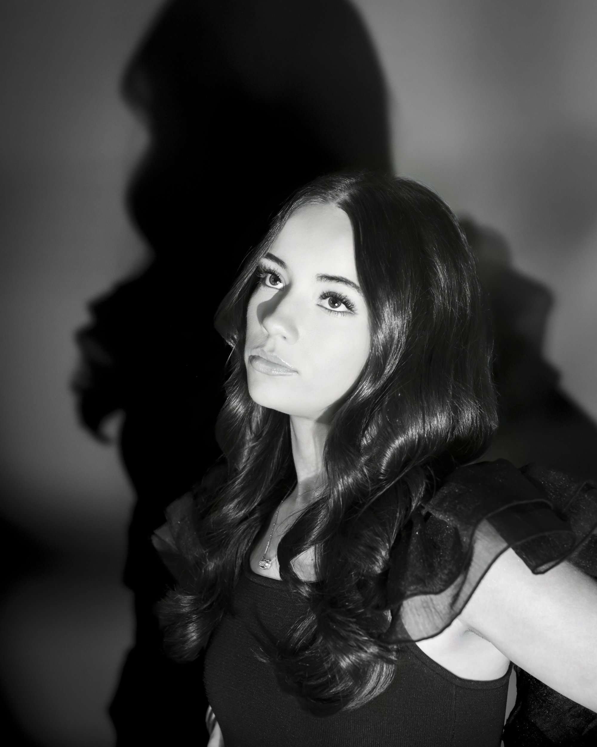 Black and white portrait of a teenage girl with long dark hair looking upward, her shadow cast on the backdrop