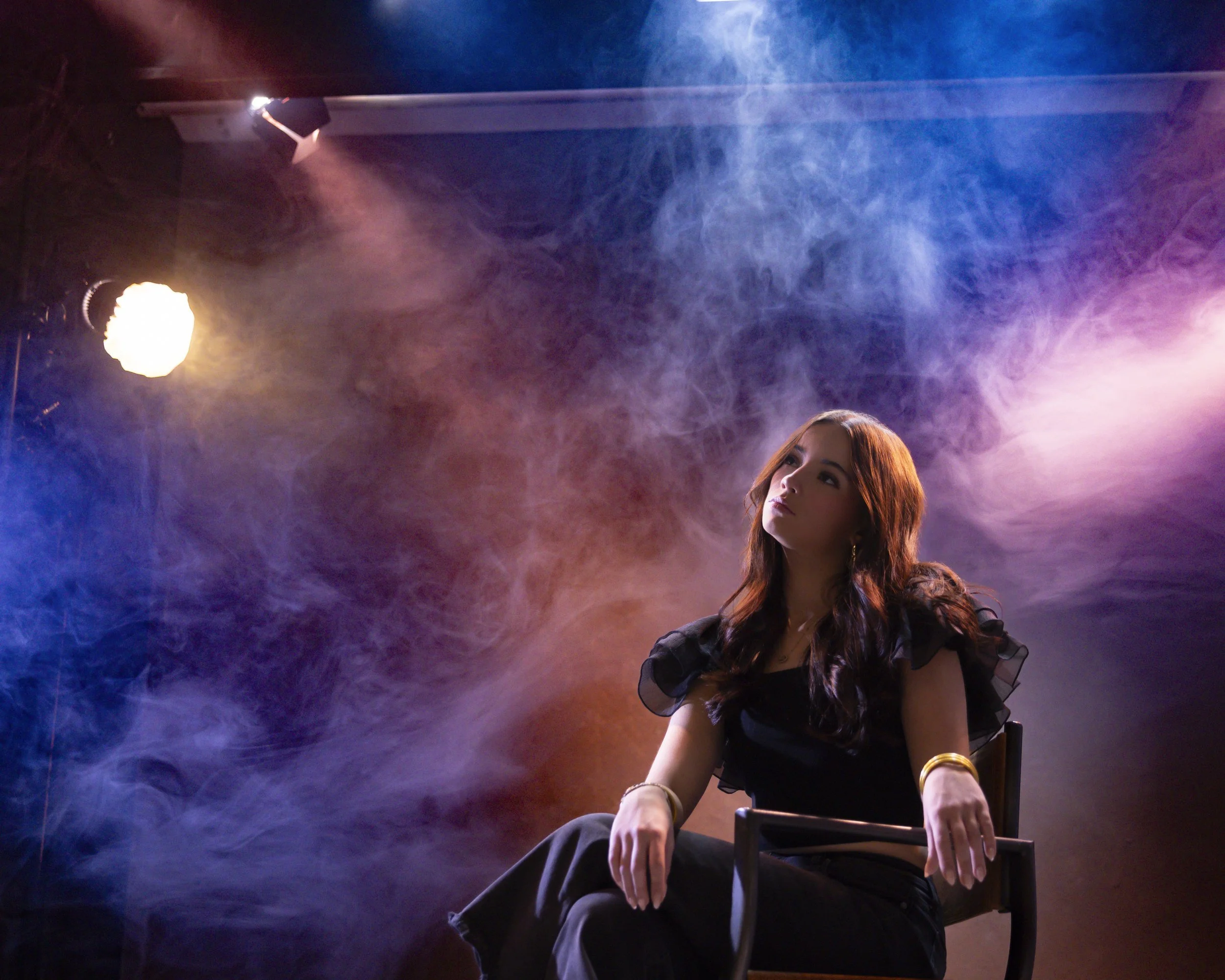 Teenage girl seated in blue and purple stage smoke and spotlights, looking upward