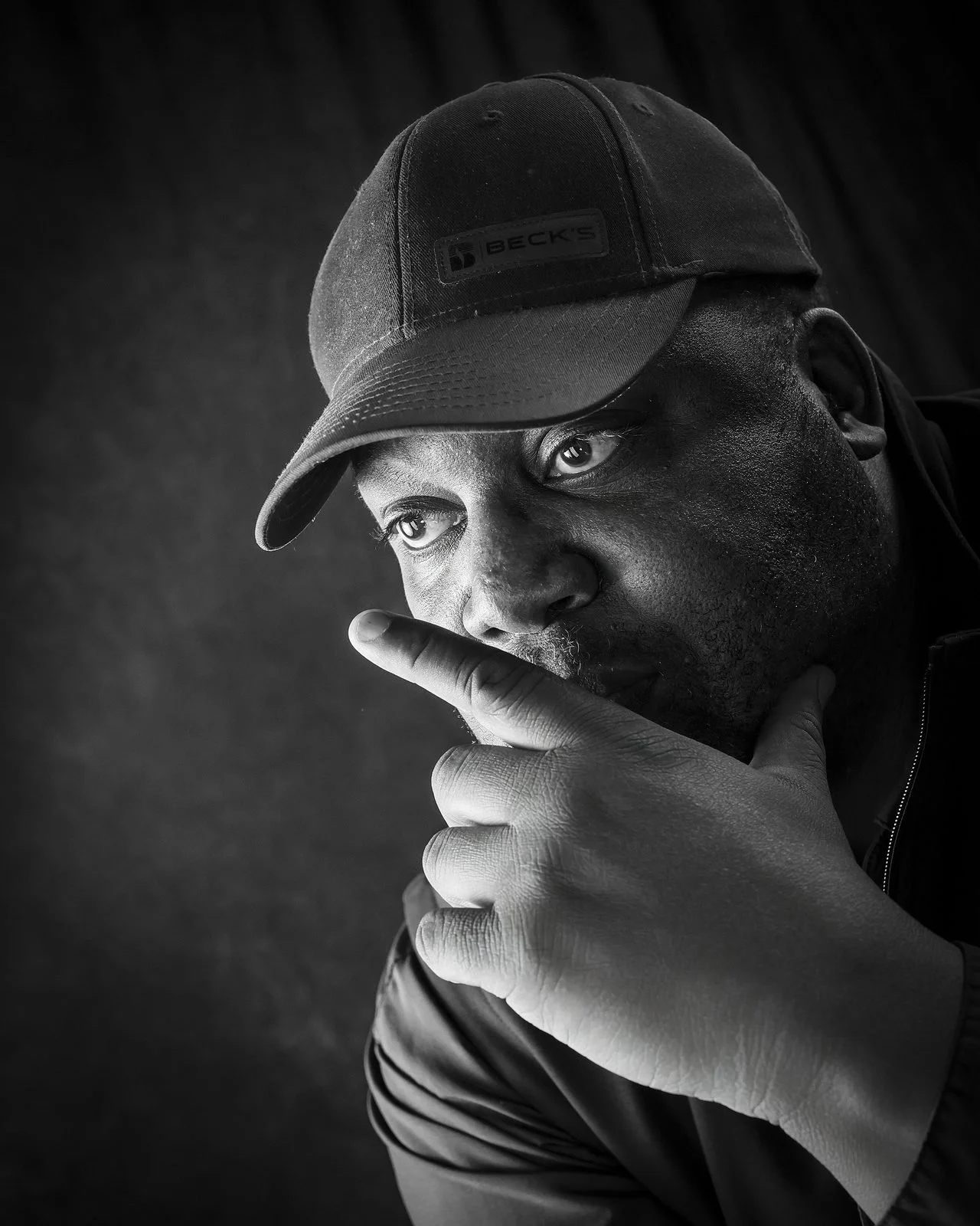 Black and white portrait of a man in a cap with his hand near his face, looking thoughtfully at the camera