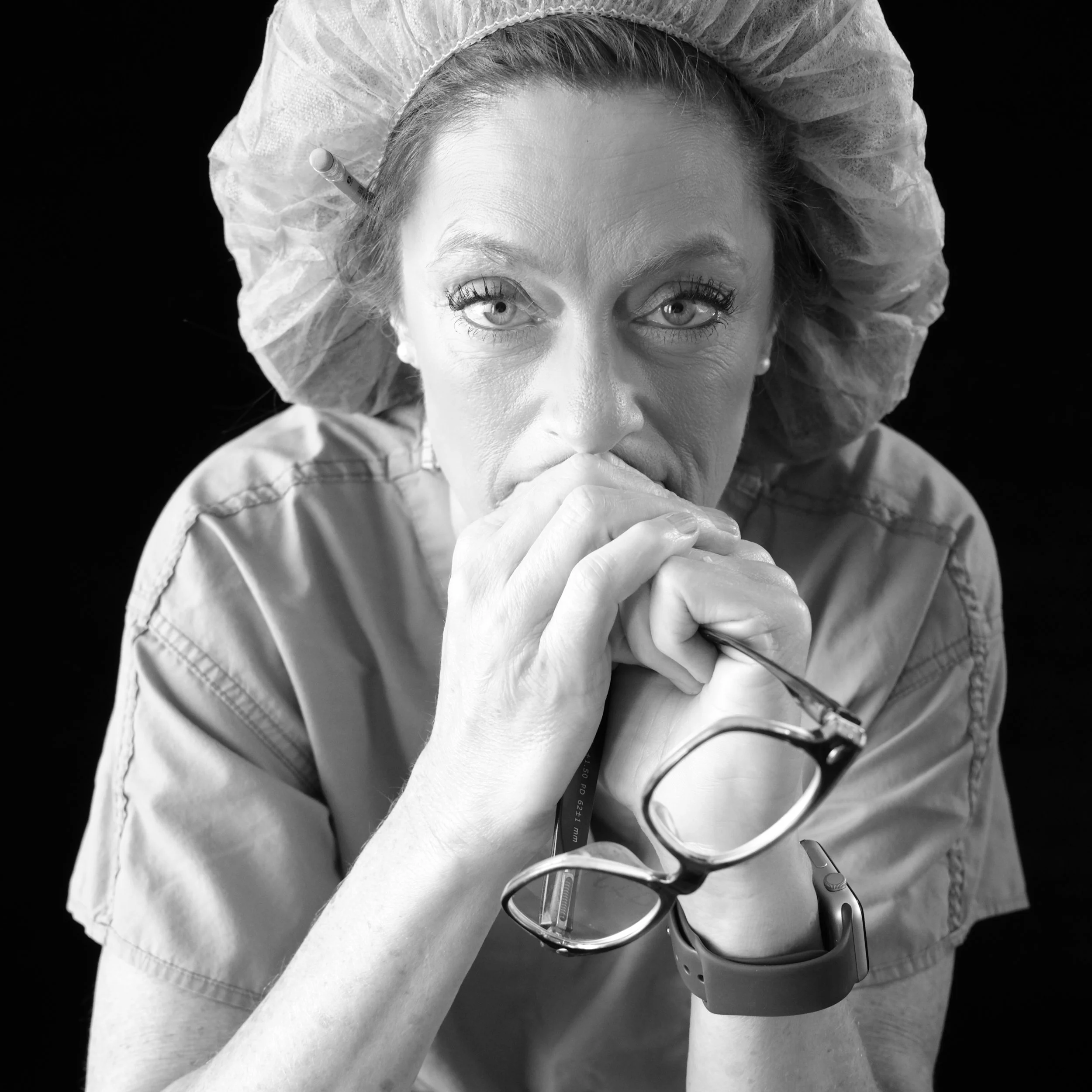Black and white portrait of a healthcare worker in scrubs and a surgical cap, hands clasped at her mouth, glasses in hand