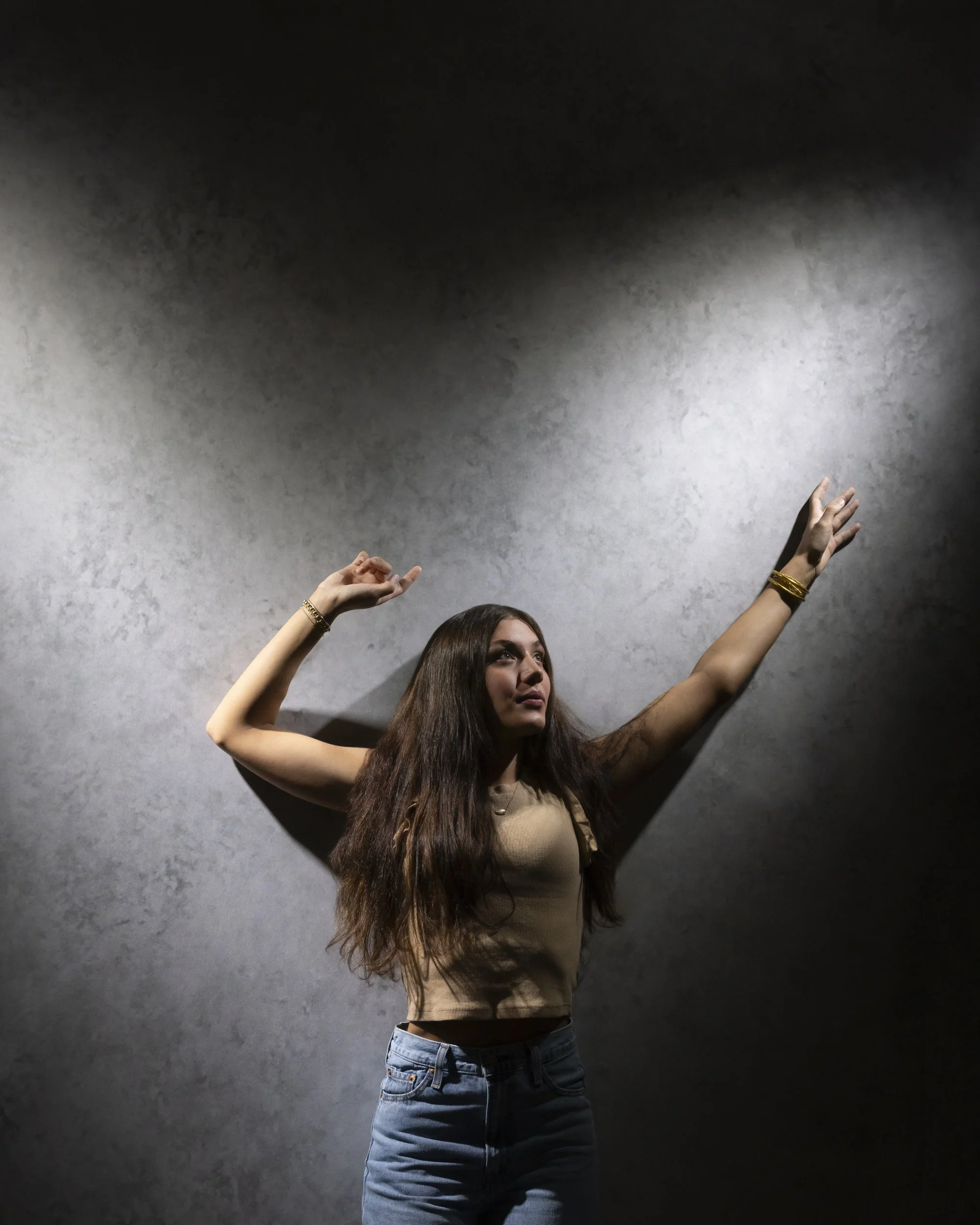Teenage girl dancing against a gray wall with her arms raised, in a beige top and blue jeans