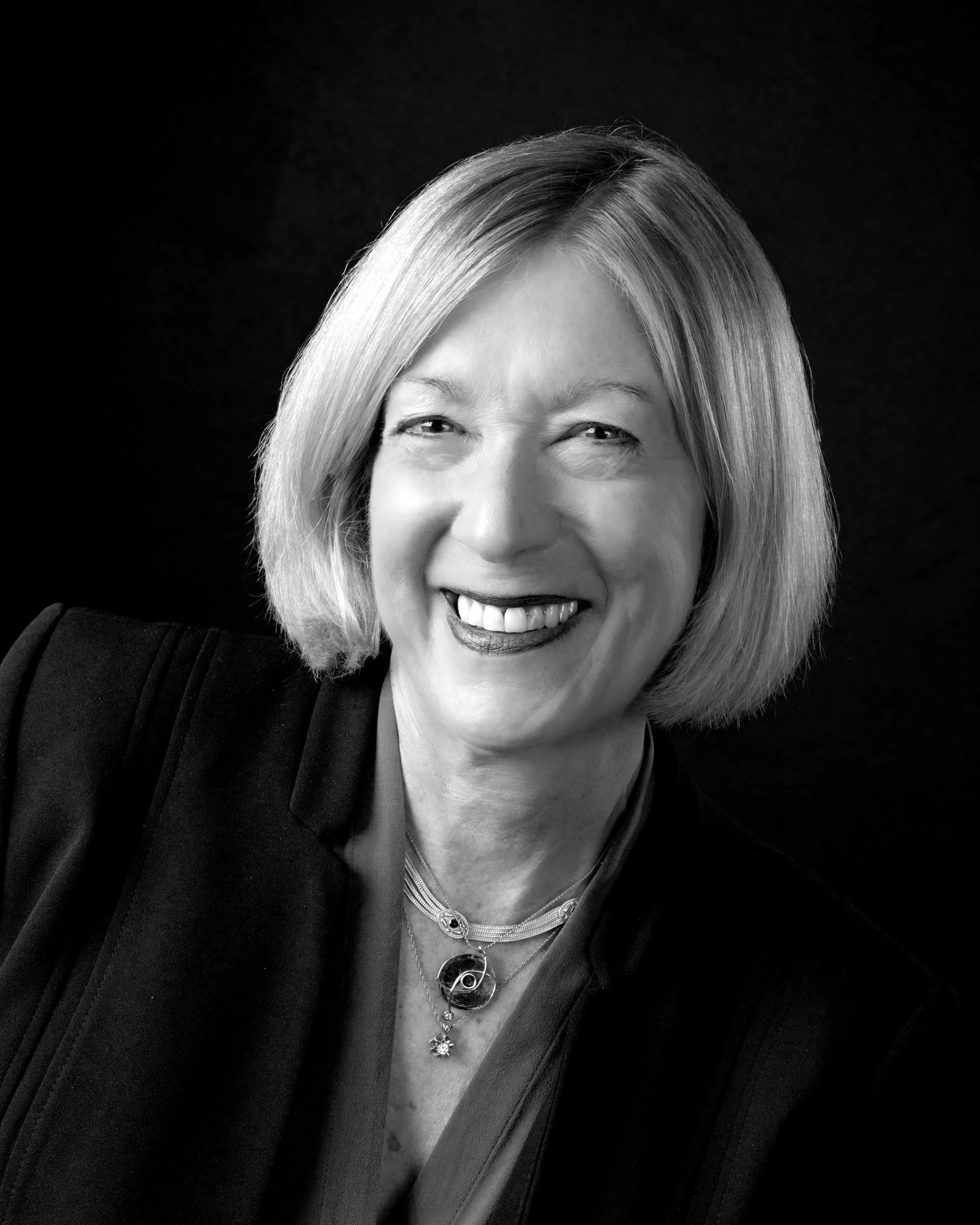 Professional black and white headshot of a smiling woman with shoulder-length hair in a dark blazer and layered necklaces