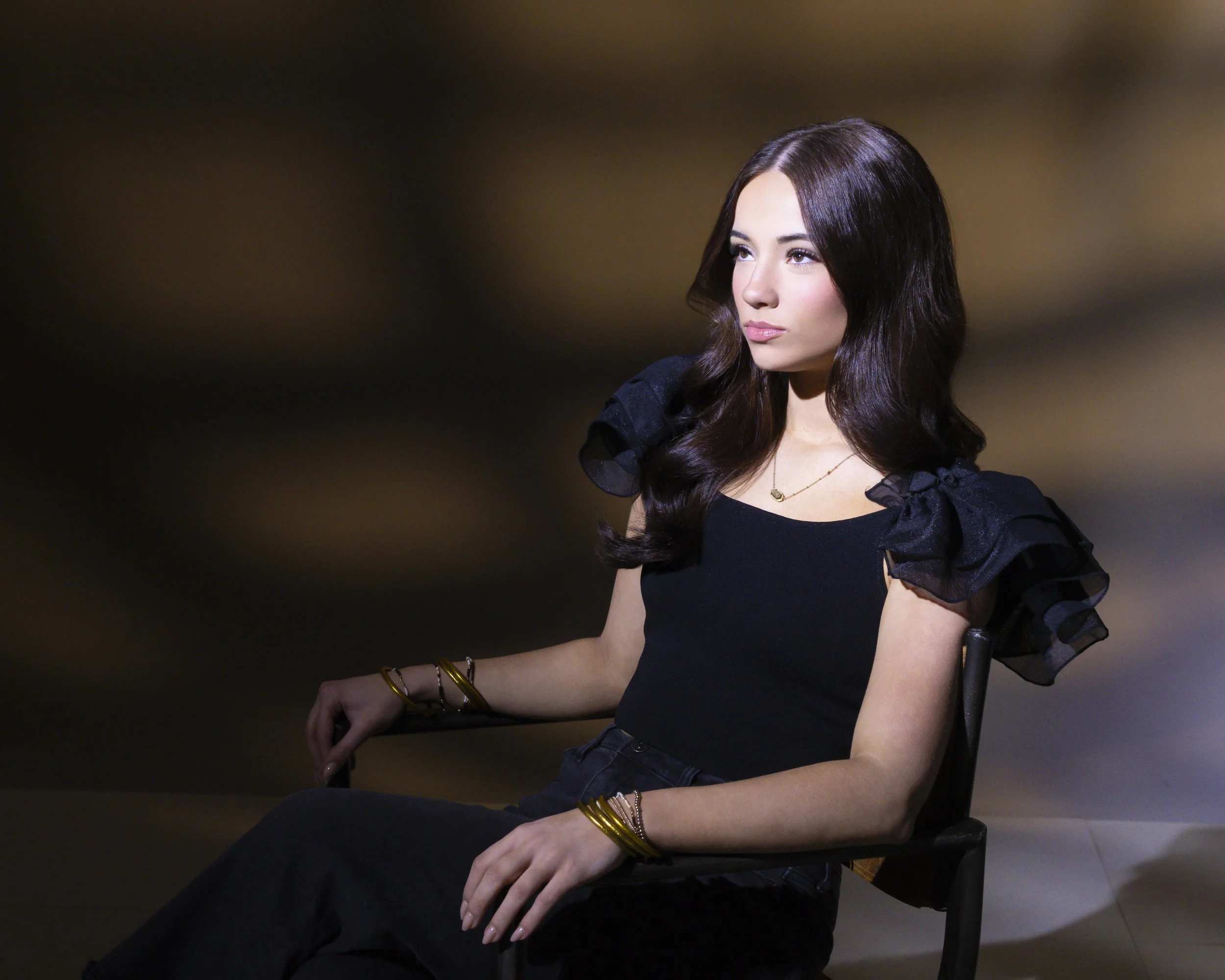 Teenage girl with long dark hair seated in a black chair, wearing a black top with ruffled sleeves and gold jewelry