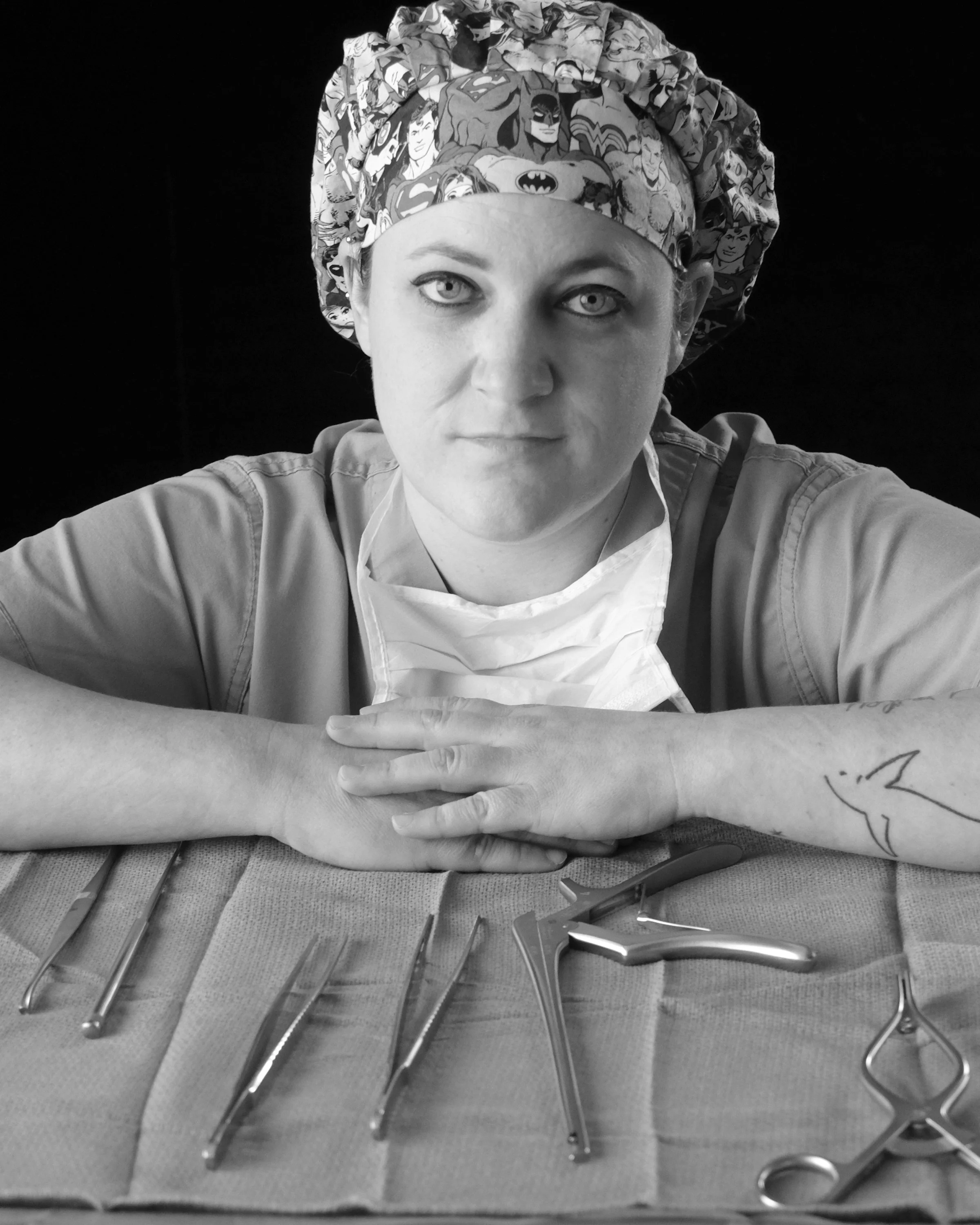 Black and white portrait of a healthcare worker in a patterned scrub cap, arms folded on a table of surgical instruments