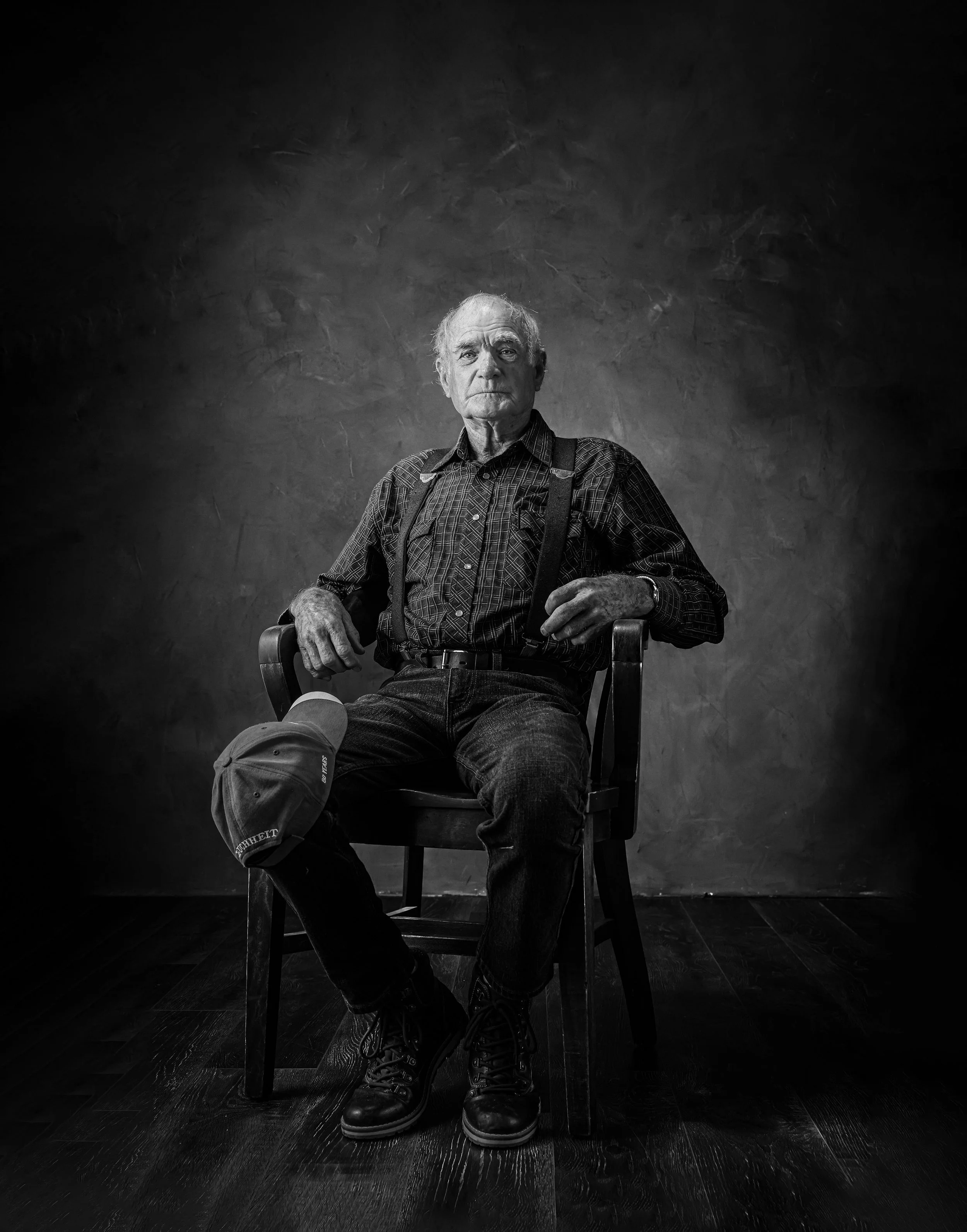 Black and white portrait of an elderly farmer seated in a wooden chair, a cap resting on his knee