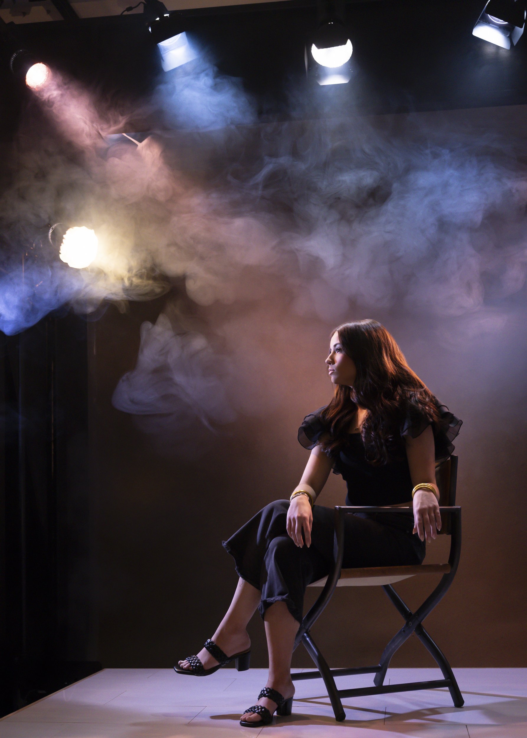 Teenage girl seated on a director-style chair in colored stage lights and smoke, in a black dress and heels
