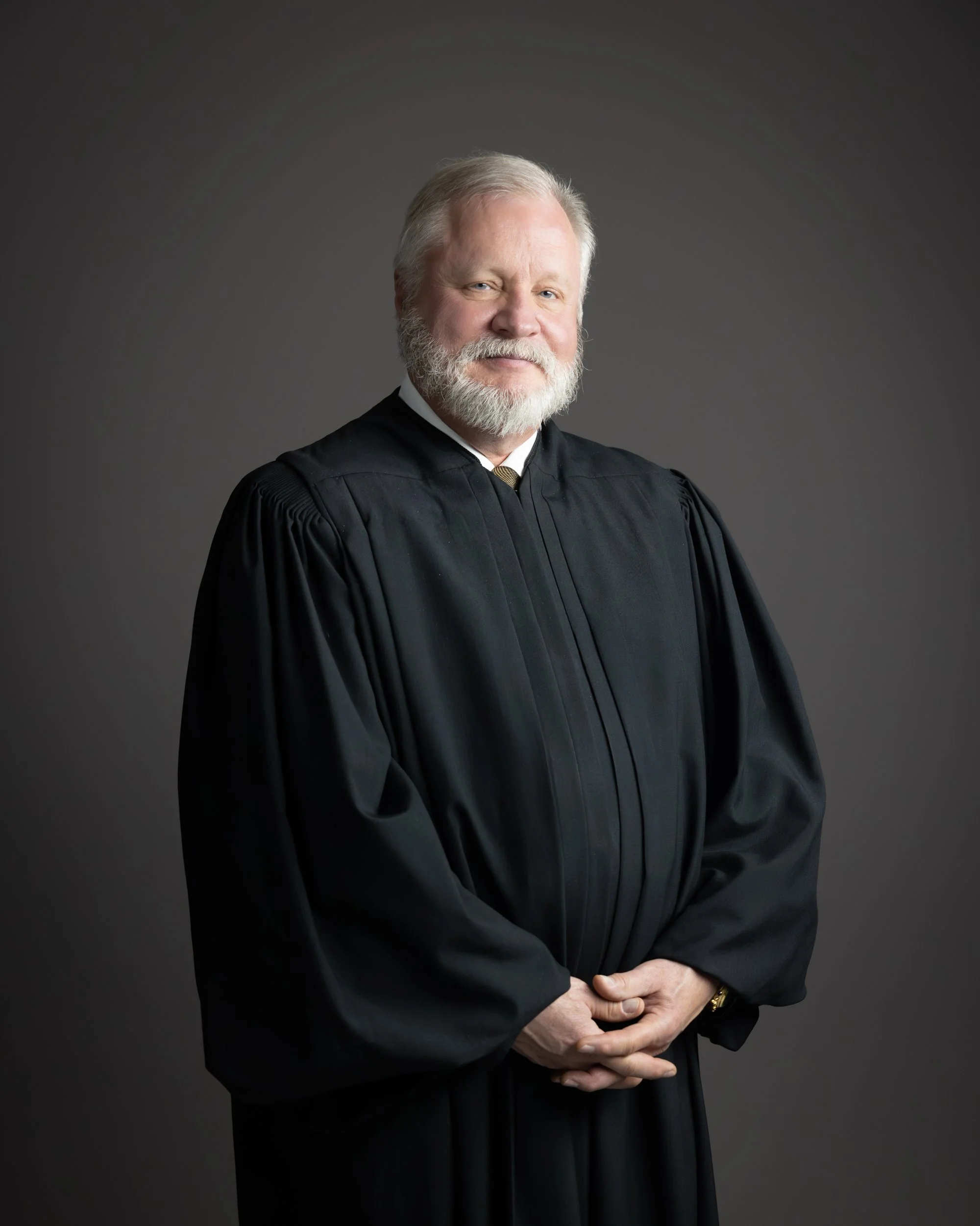 Professional portrait of a gray-haired, bearded man in a judge's black robe against a dark gray background