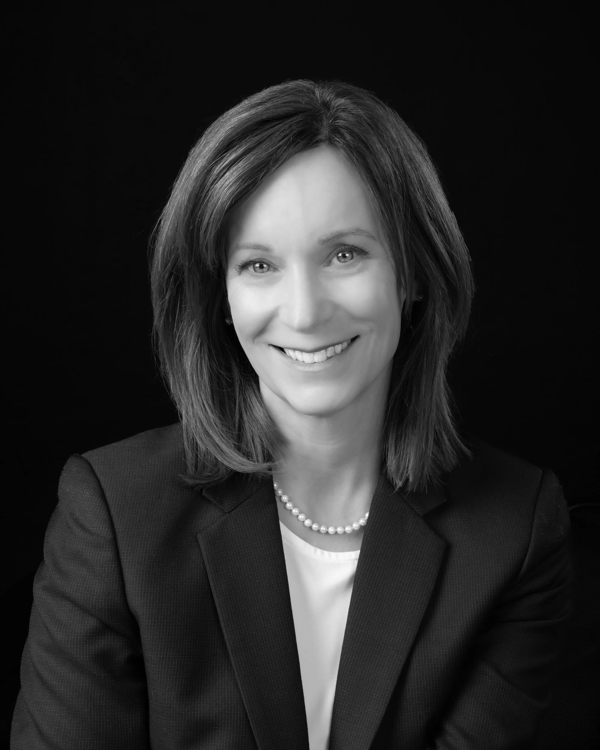 Smiling professional black and white headshot of a woman in a dark blazer with a pearl necklace