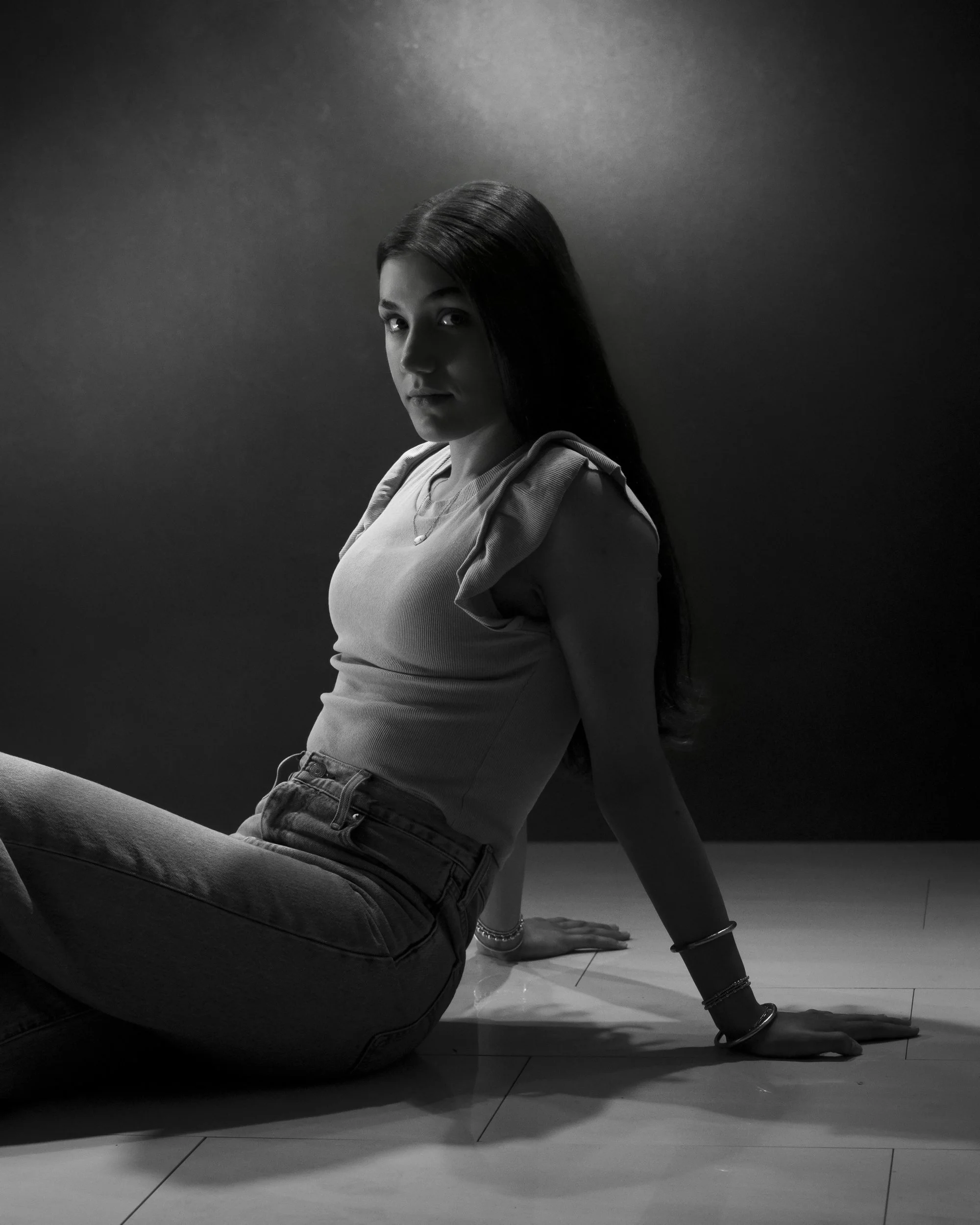 Black and white portrait of a teenage girl seated on the floor, leaning back on one arm and looking at the camera