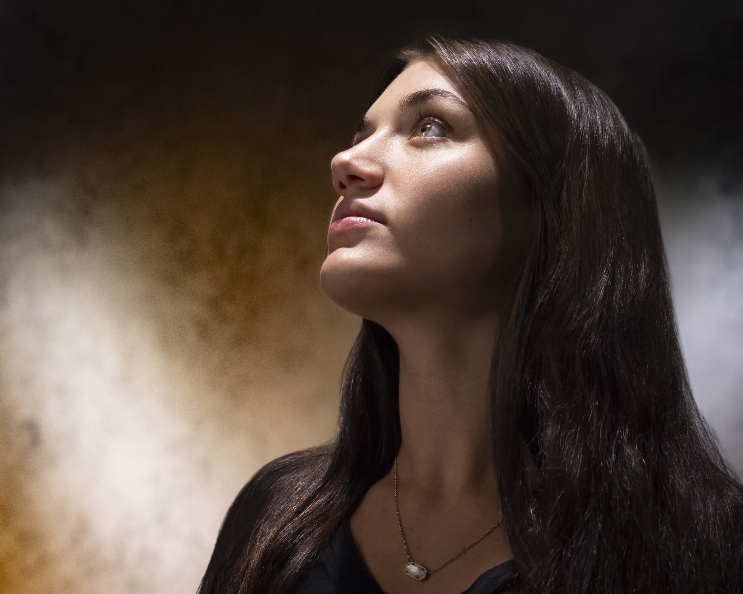 Close portrait of a teenage girl with long dark hair looking upward against a softly blurred golden background