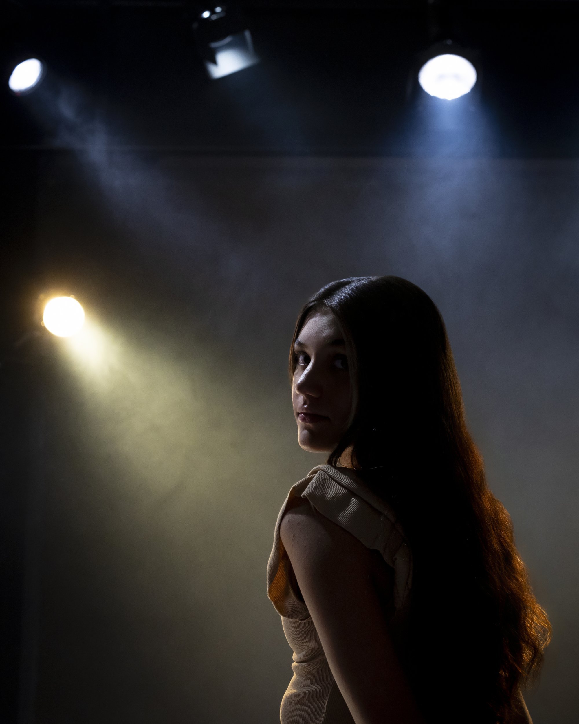 Teenage girl with long hair in a dark room lit by stage spotlights, looking over her shoulder