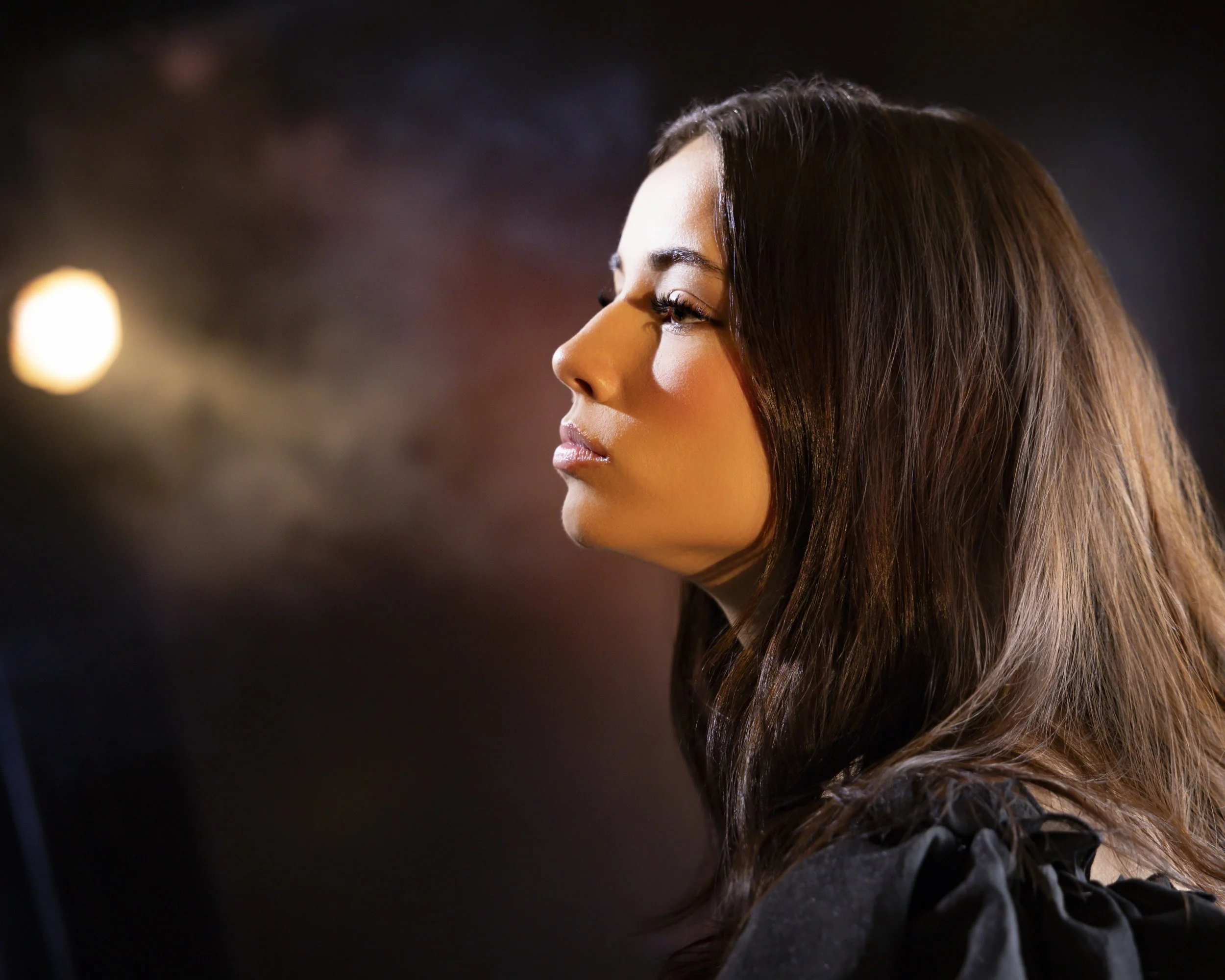 Side profile of a teenage girl with long brown hair in dark clothing against soft, blurred lighting