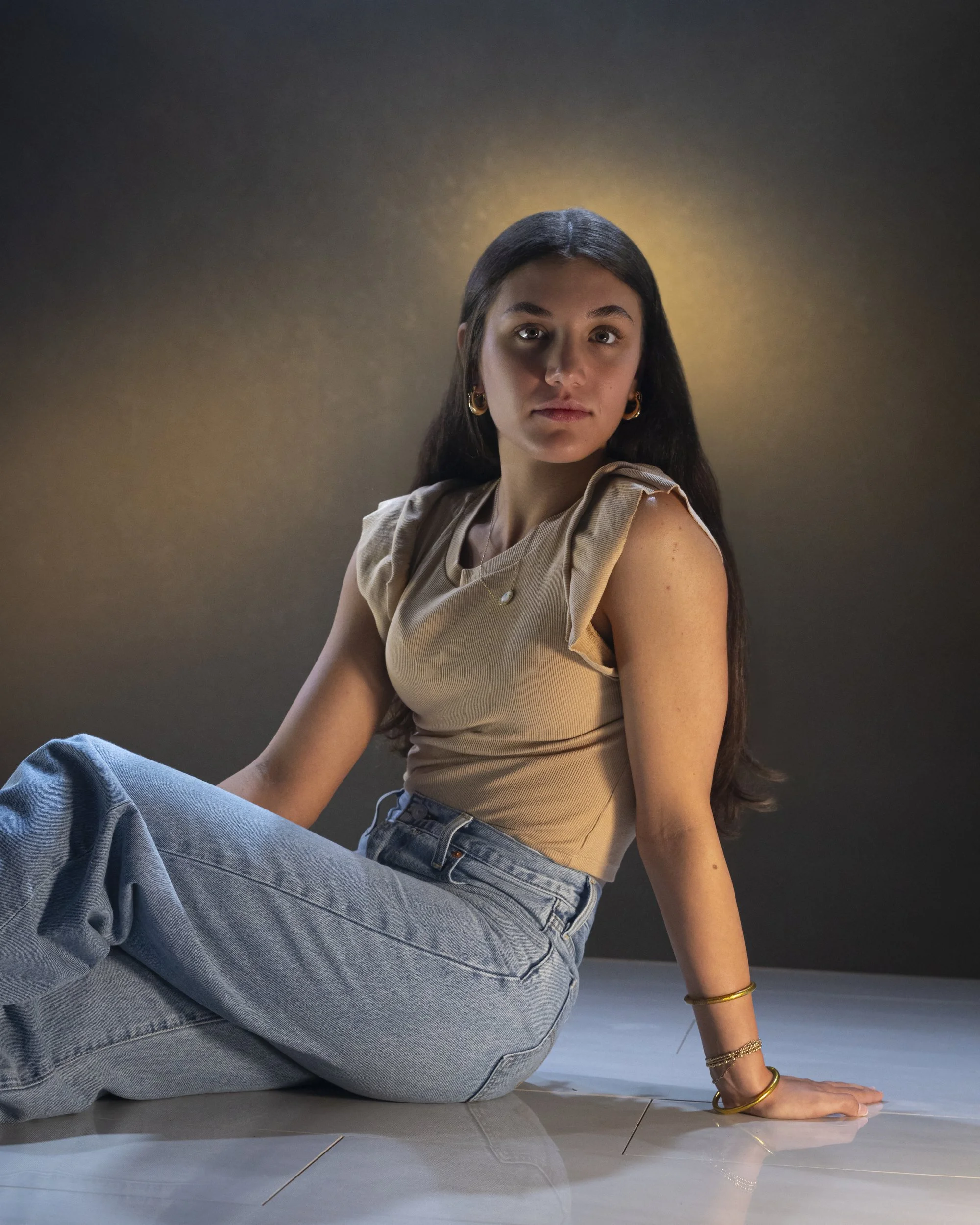Teenage girl in a beige top and jeans seated on the floor in warm golden light against a dark backdrop