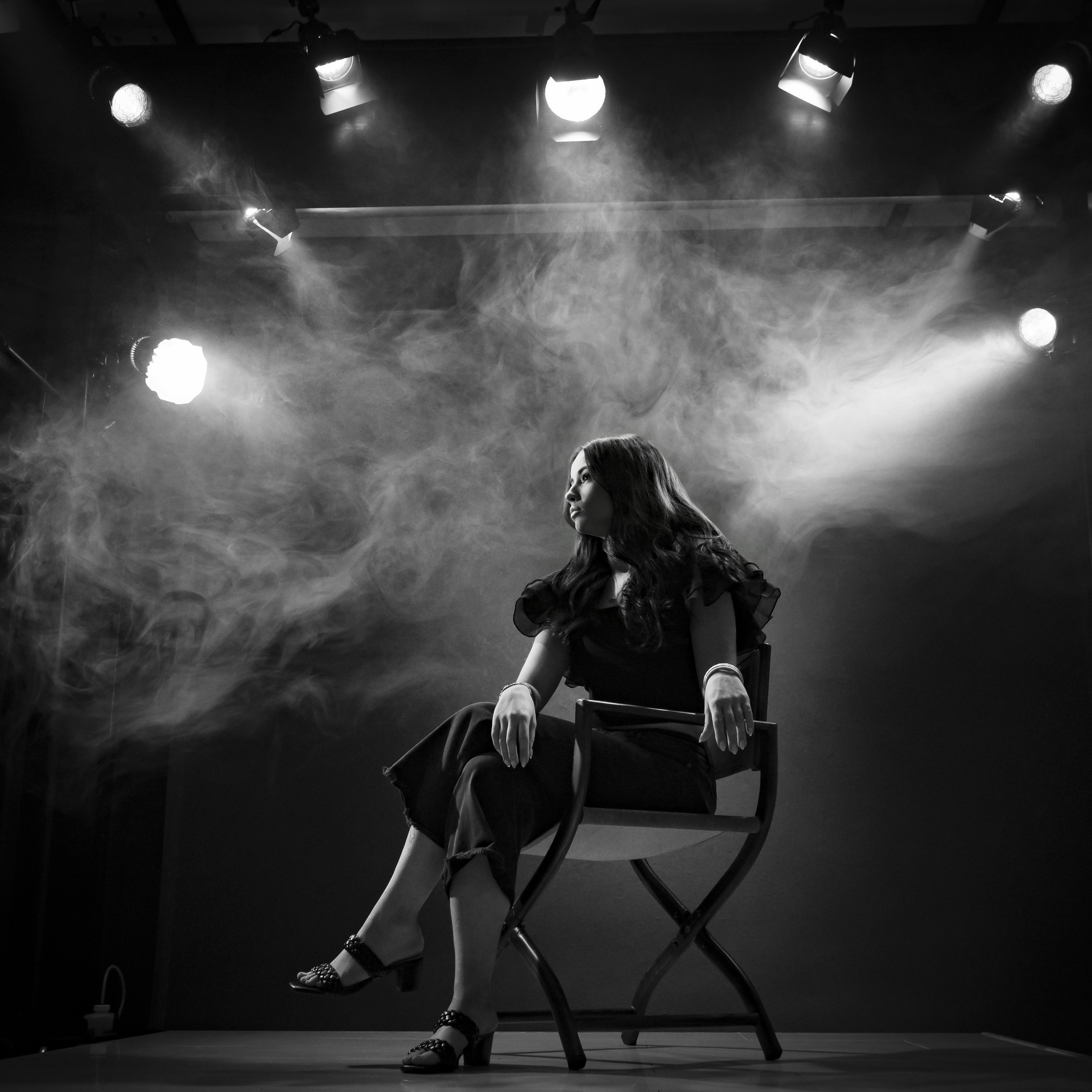 Black and white portrait of a teenage girl seated on a chair with crossed legs under stage lights and haze