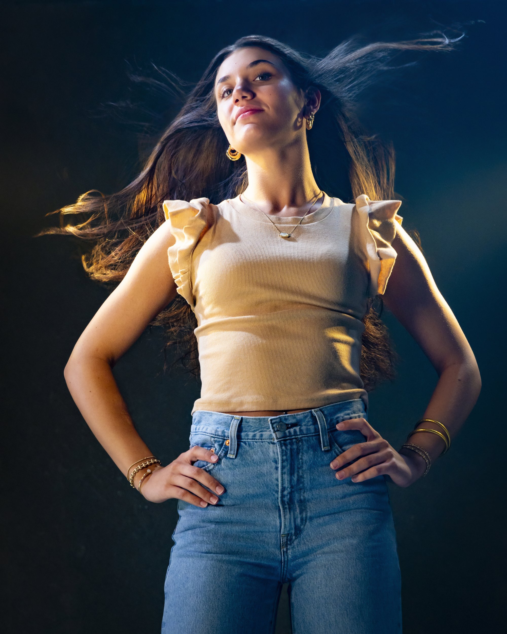 Teenage girl standing with her hands on her hips in a beige top and jeans, long hair flowing, against a dark background