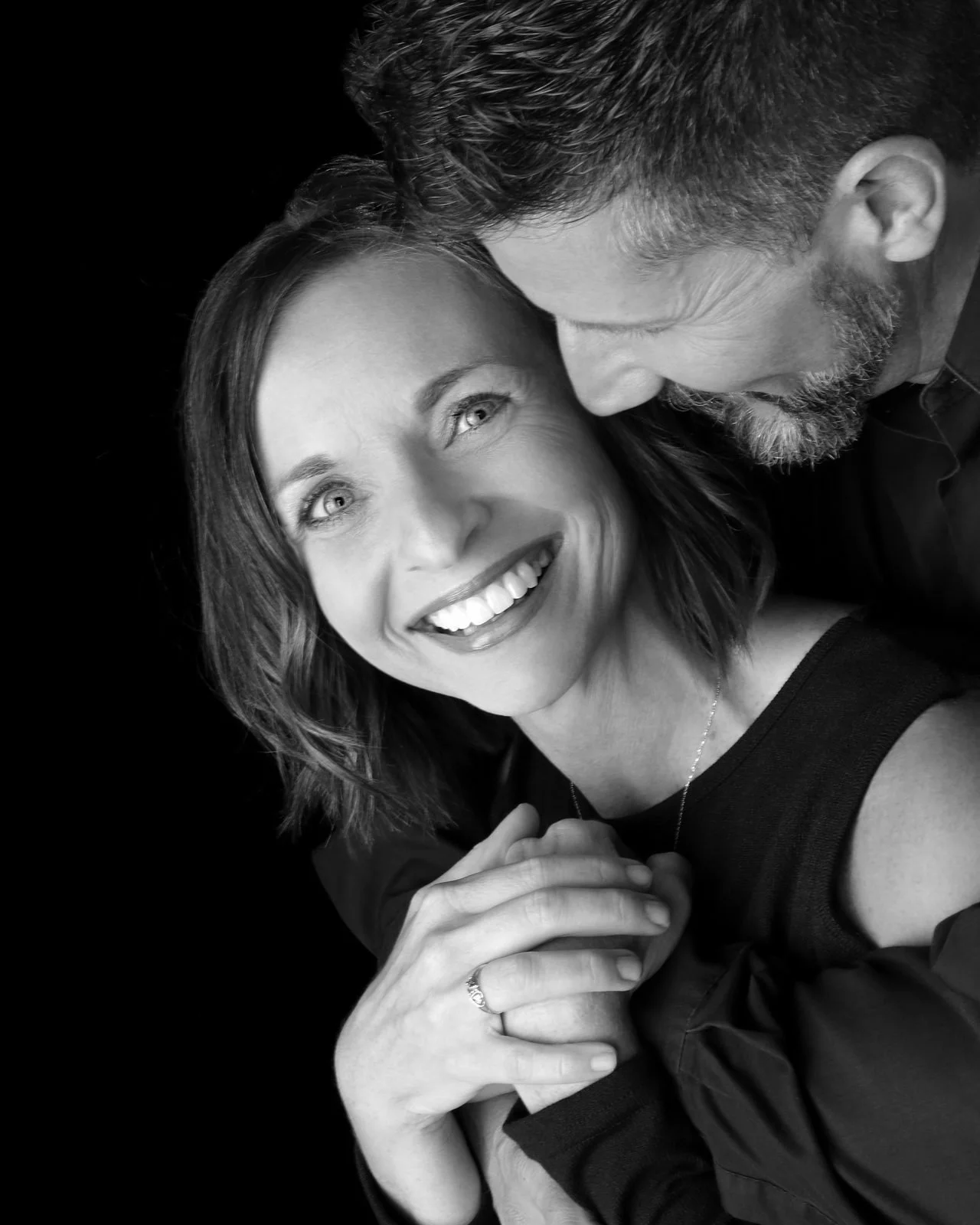 Engaged couple, a smiling woman showing her ring as her partner rests his forehead on her temple, black and white portrait