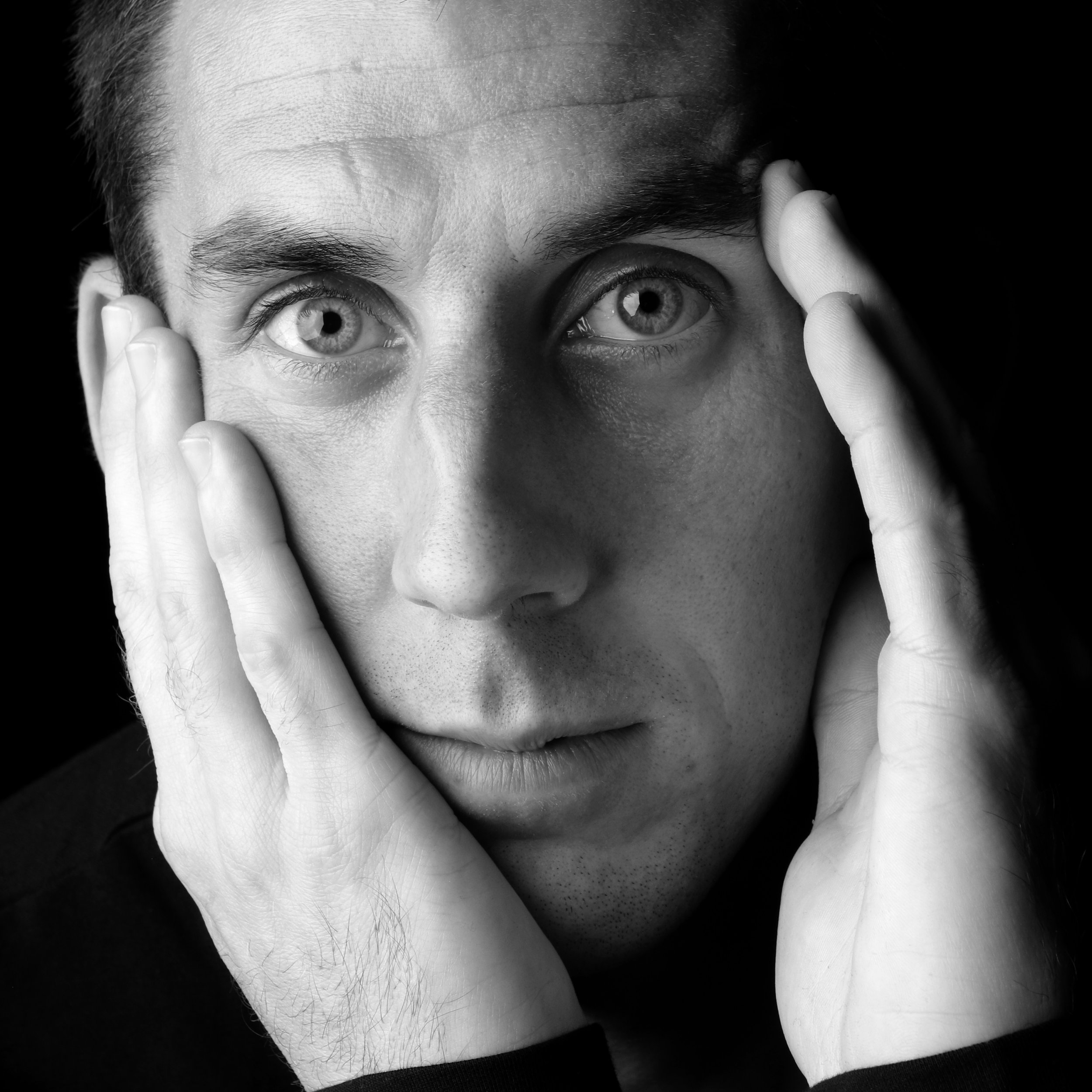 Close black and white portrait of a man with both hands pressed to his face, looking straight into the camera