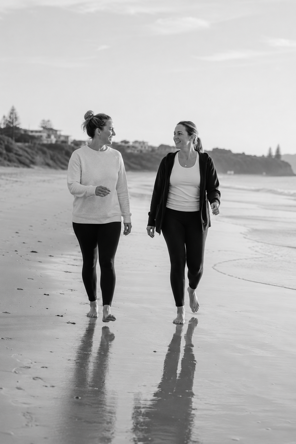 Two women walking barefoot on a beach and talking, with houses and cliffs in the background.