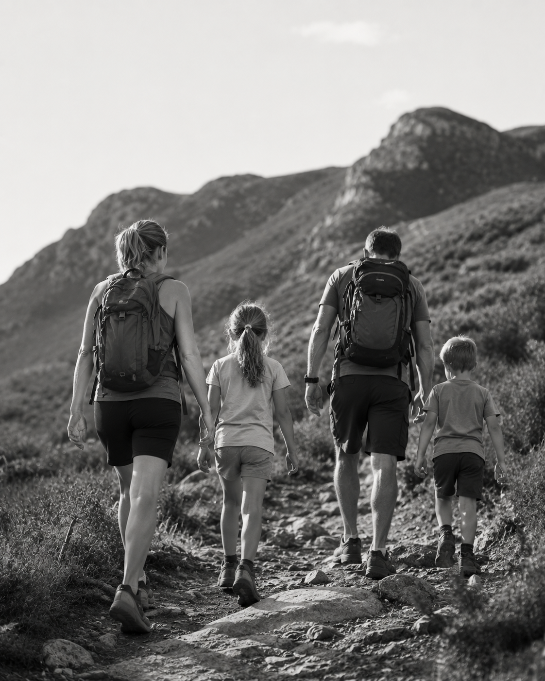A family of four hiking up a rocky trail through a mountainous landscape, all carrying backpacks. The photo is in black and white.