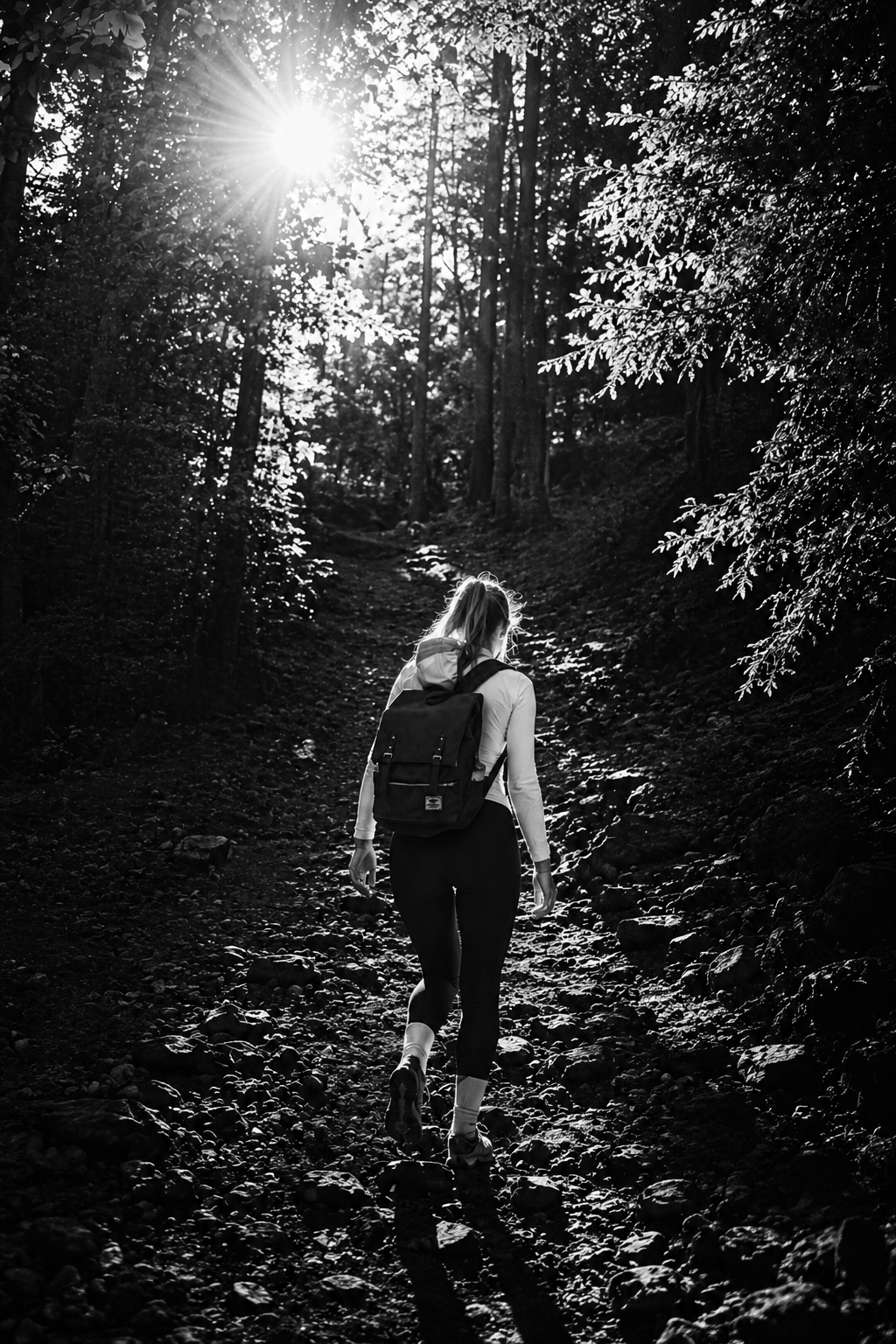 A woman hiking on a rocky forest trail during daytime with sunlight shining through trees.