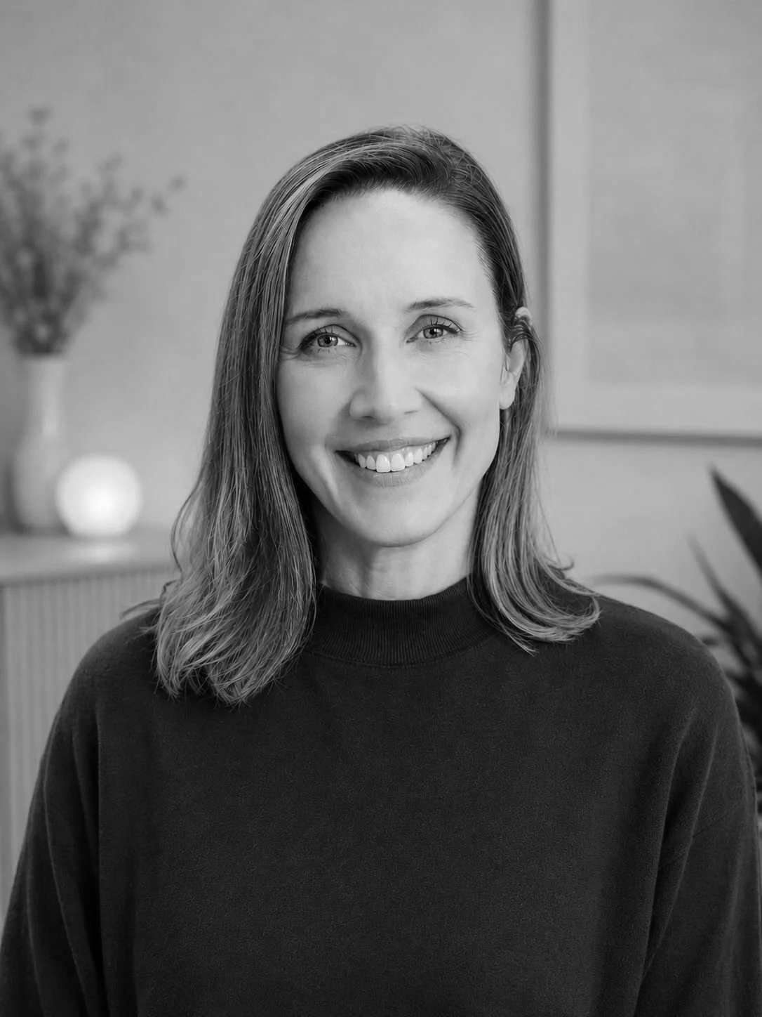 Black and white portrait of a smiling woman with shoulder-length hair, wearing a dark top, indoors with blurred background including a vase with flowers and a framed picture.